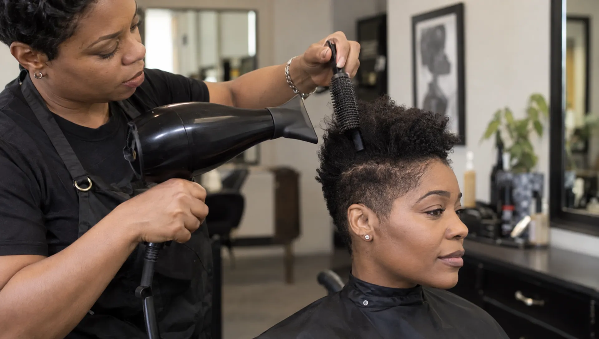A stylist uses a concentrator nozzle and small styling brush on short natural hair, stretching the tapered top while keeping the sides close.
