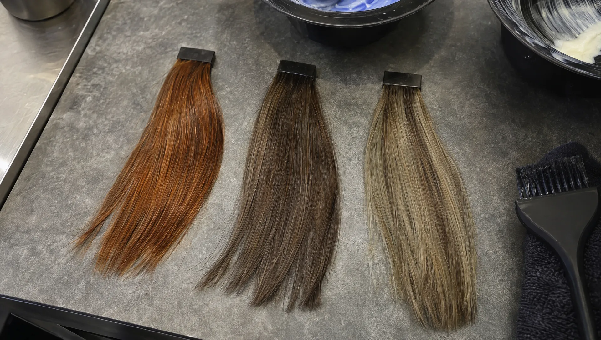 A close overhead view of three brunette hair swatches on a salon colorist's work surface: one copper-orange, one cooler brown after blue shampoo, and one pale highlighted swatch with a greenish cast.