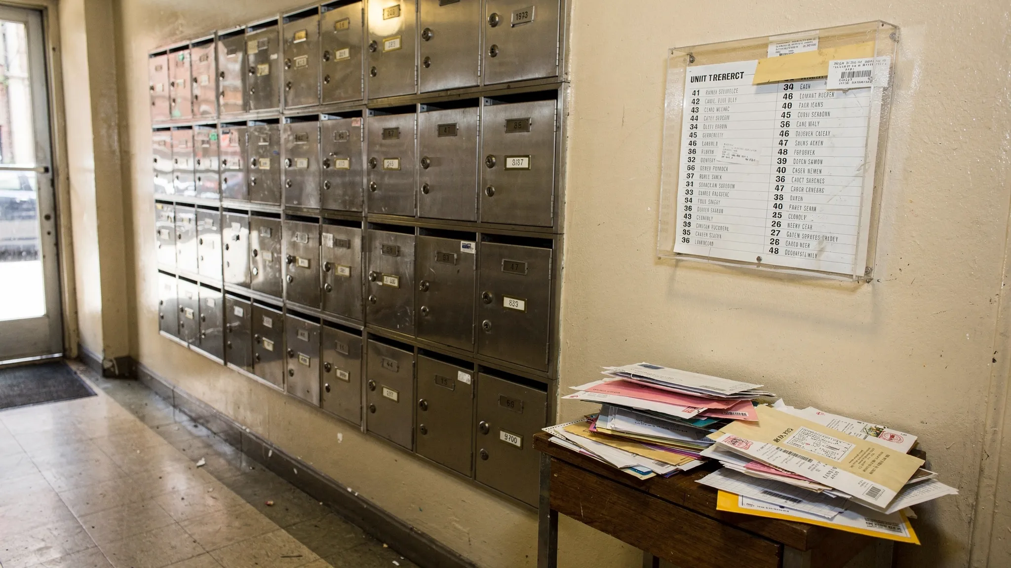 A typical NYC small-building mailbox wall with clearly labeled unit mail slots, a simple lobby directory listing unit numbers and names, and a returned-mail pile on a small table nearby.