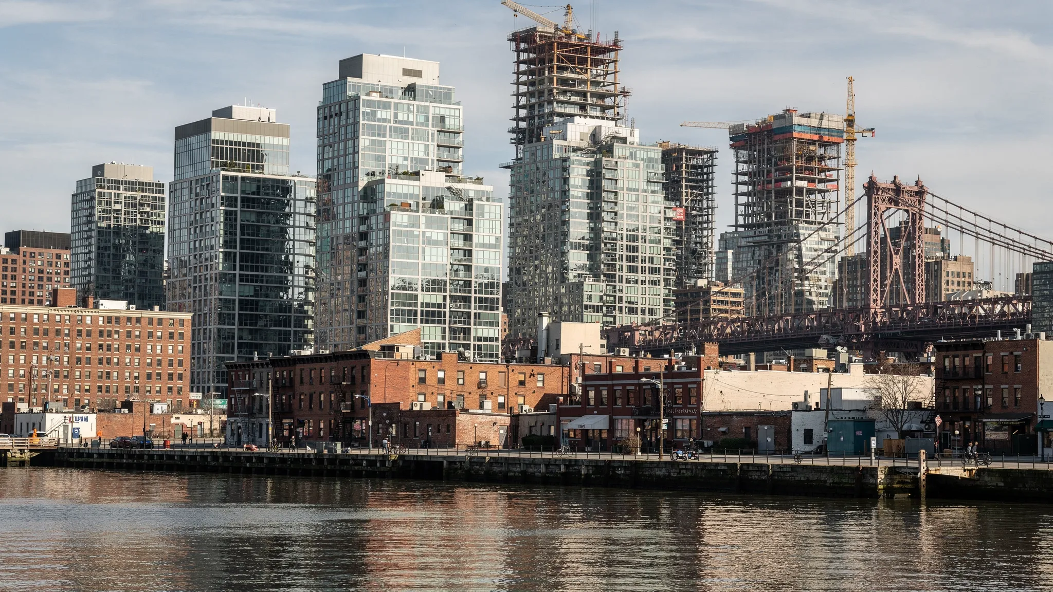 A street-level view of Long Island City’s waterfront skyline with a mix of modern condo towers and older low-rise buildings, showing construction activity in the background and the Queensboro Bridge in the distance.