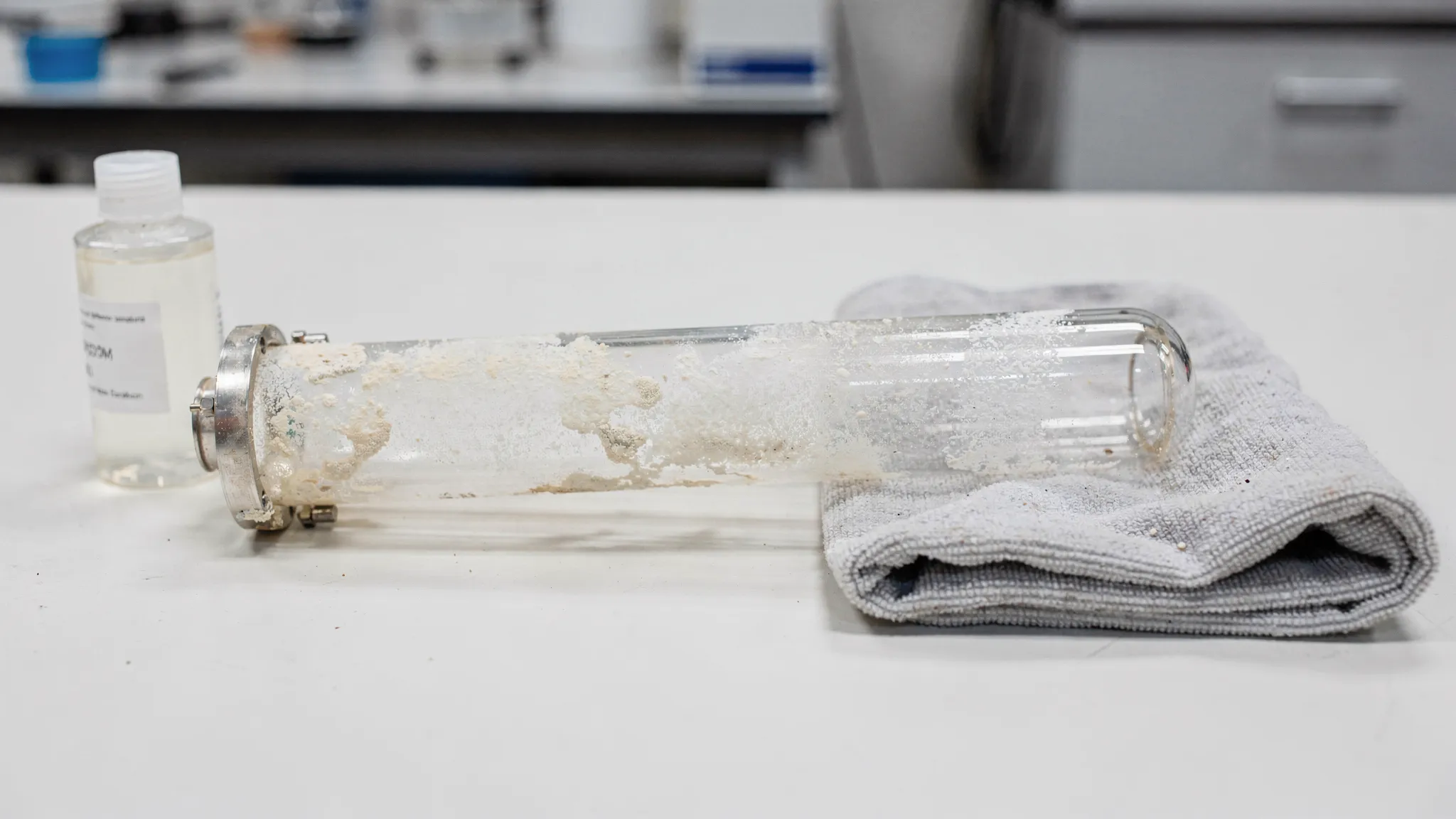 Un tub de cuarț (Quartz UV) scos din camera unui sterilizator UV, cu depuneri vizibile de calcar și zone mai transparente, alături de o lavetă moale și un recipient cu soluție de curățare, pe o masă de lucru curată.