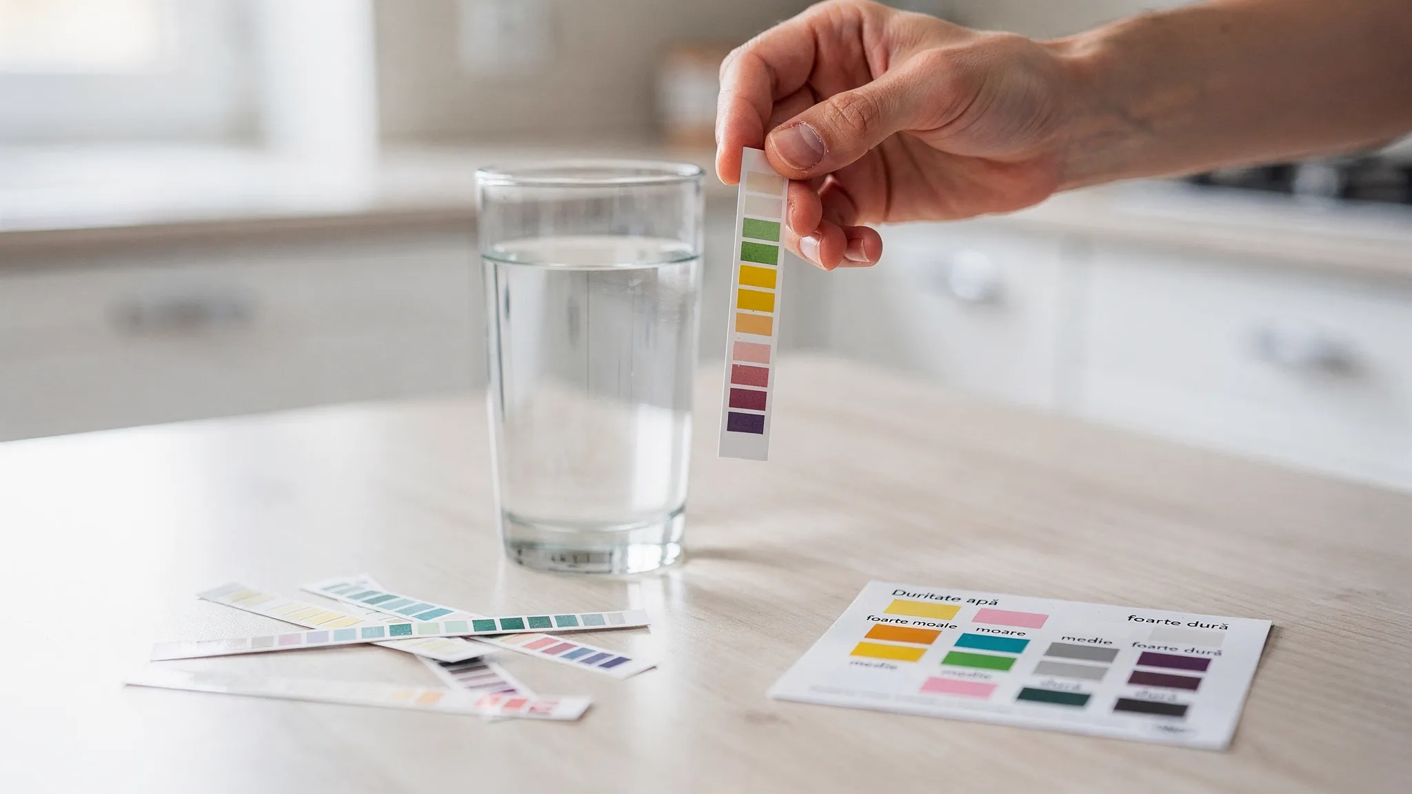 O persoană ține în mână o bandă de test pentru duritatea apei lângă un pahar transparent cu apă, pe masă se văd câteva benzi suplimentare și o mică fișă de interpretare a culorilor.