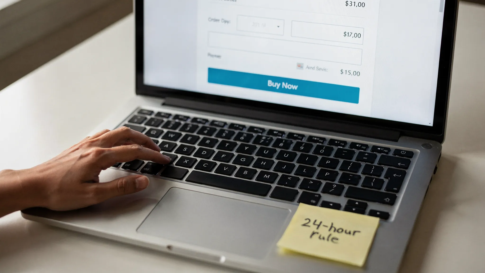 Close-up of a laptop checkout page showing a prominent âBuy Nowâ button. The shopperâs hand hovers over the trackpad while a sticky note on the keyboard reads â24-hour rule.â The browserâs saved payment options are removed.