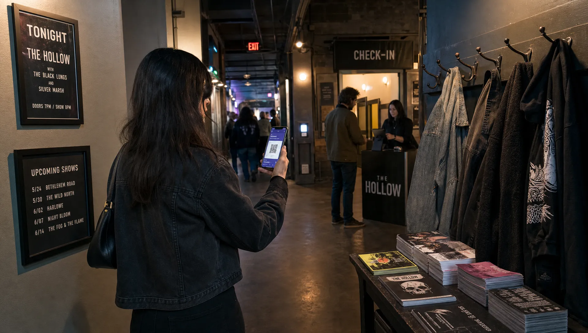 Landscape medium scene in a music venue corridor during opening time, where an attendee pulls up a digital ticket on a phone beside a coat rack, a stacked flyer table, and a staff check-in station deeper in the hall. The moment should feel like ordinary arrival behavior before the line reaches the scanner, with the venue interior doing the storytelling.