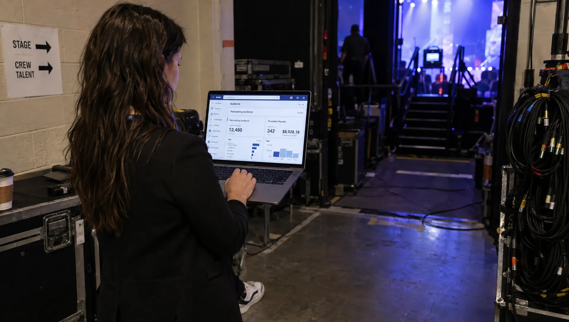 Landscape over-the-shoulder view of an event marketer standing at a venue side wall, using a laptop with the screen facing the camera to review a Facebook retargeting campaign audience and purchase results. One hand rests on the trackpad; a stage entrance and coiled audio cables sit in the background, with the full person visible.