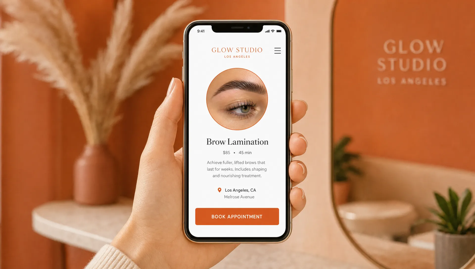 A phone held upright showing a simple booking page for a small Los Angeles beauty studio, with a clear service name, location, and appointment button visible on the screen. The phone screen faces forward and nothing appears behind the screen.