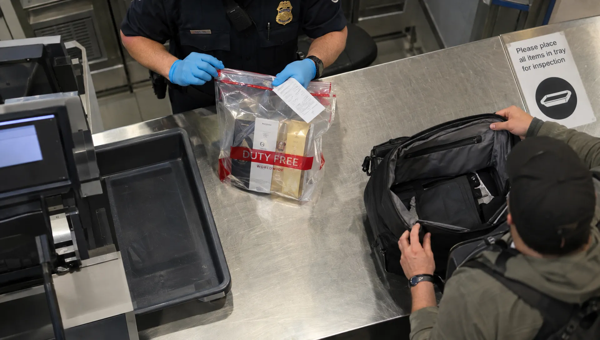 Security staff inspect a sealed duty-free toiletries bag and receipt at an airport transfer checkpoint.