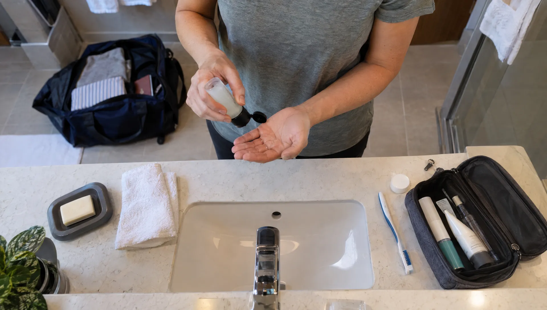 A traveler uses a small shampoo bottle at a hotel sink, with everyday toiletries and an open travel bag nearby.