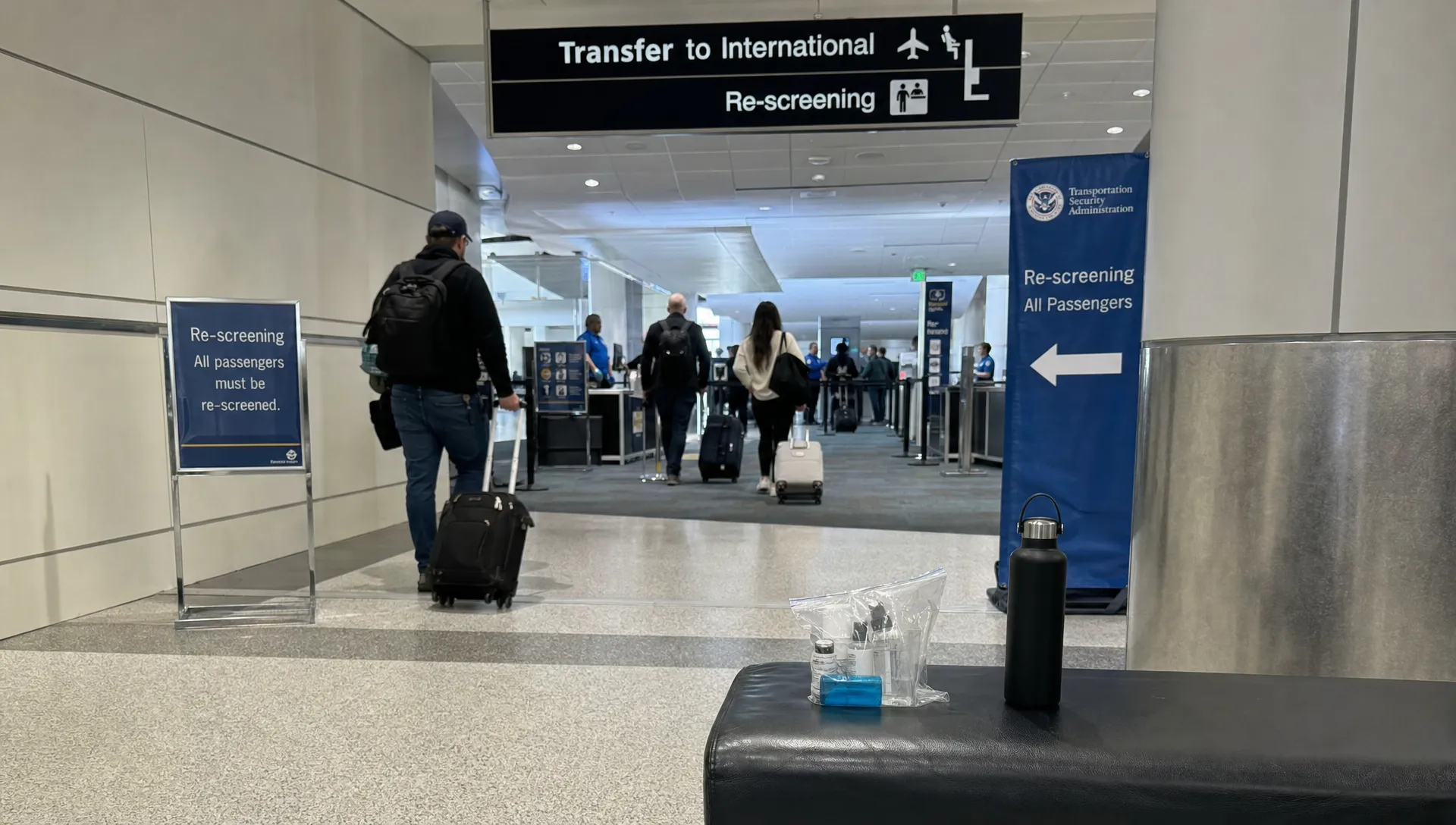 An airport transfer corridor with a re-screening checkpoint and a small liquids bag on a bench, showing how connection flights affect cabin liquid rules.