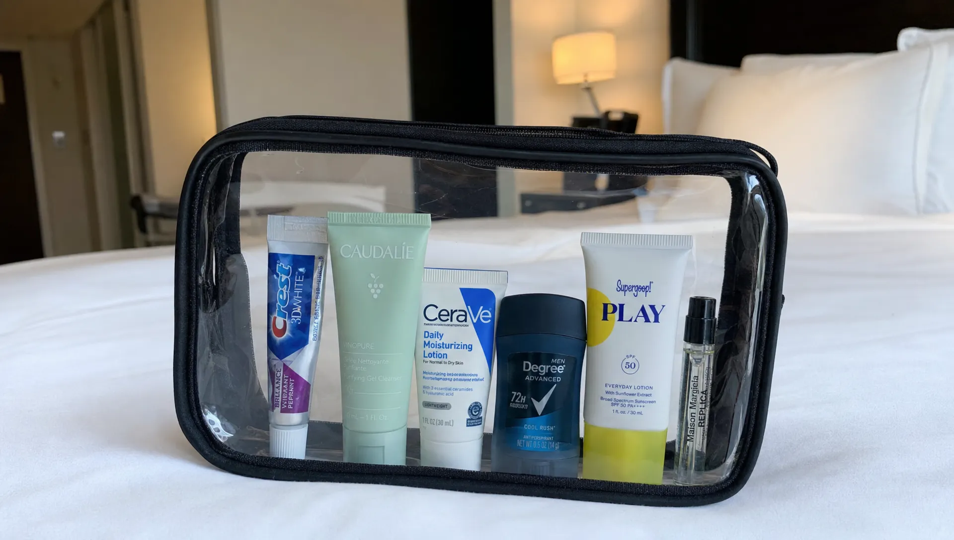 A clear toiletries bag on a hotel bed holds mini toothpaste, cleanser, moisturiser, deodorant, sunscreen and a small fragrance vial.