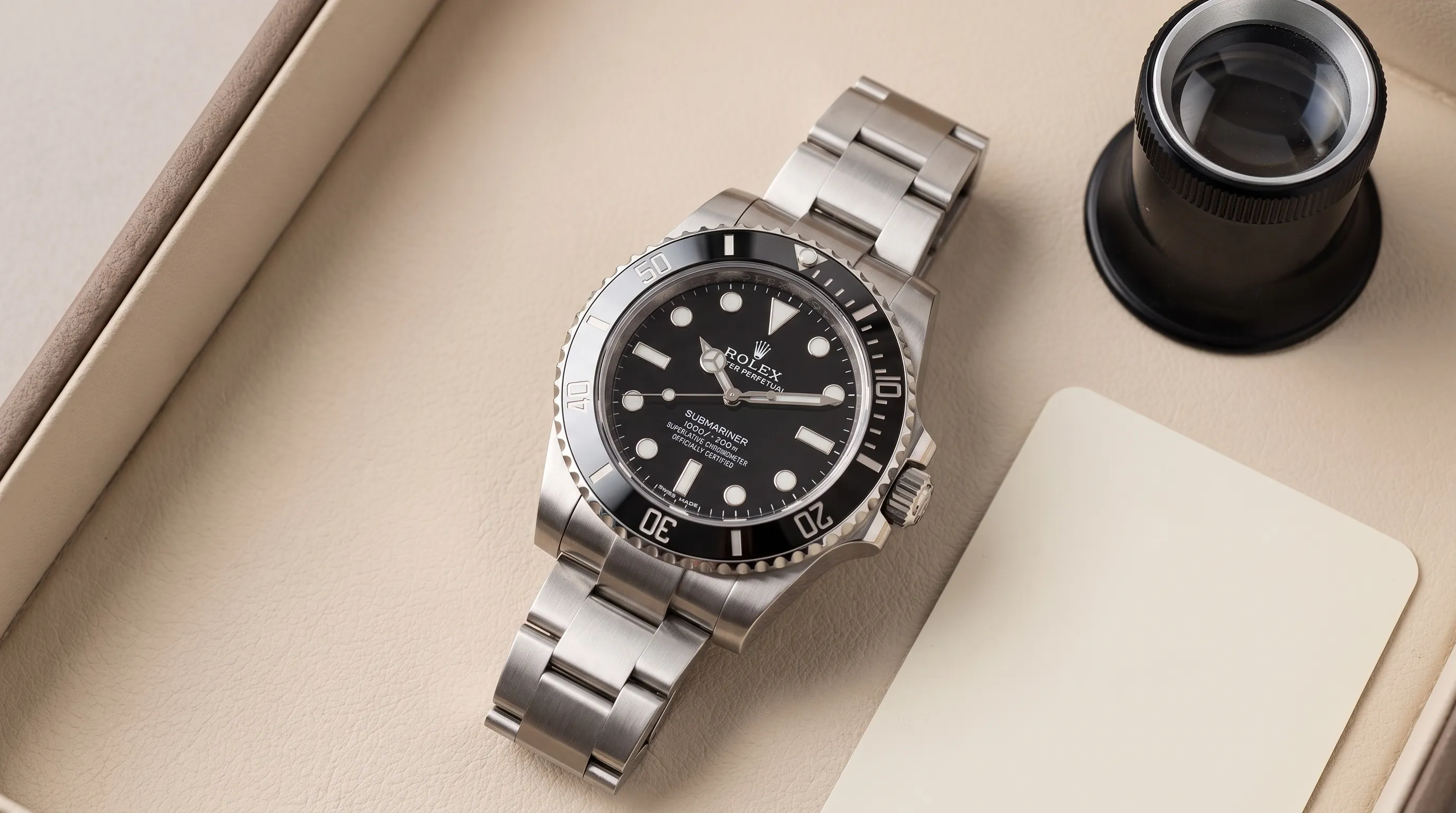 A close-up view of a stainless steel luxury wristwatch on a neutral watch tray, with a loupe and unmarked inspection card nearby to suggest careful authentication and condition review.
