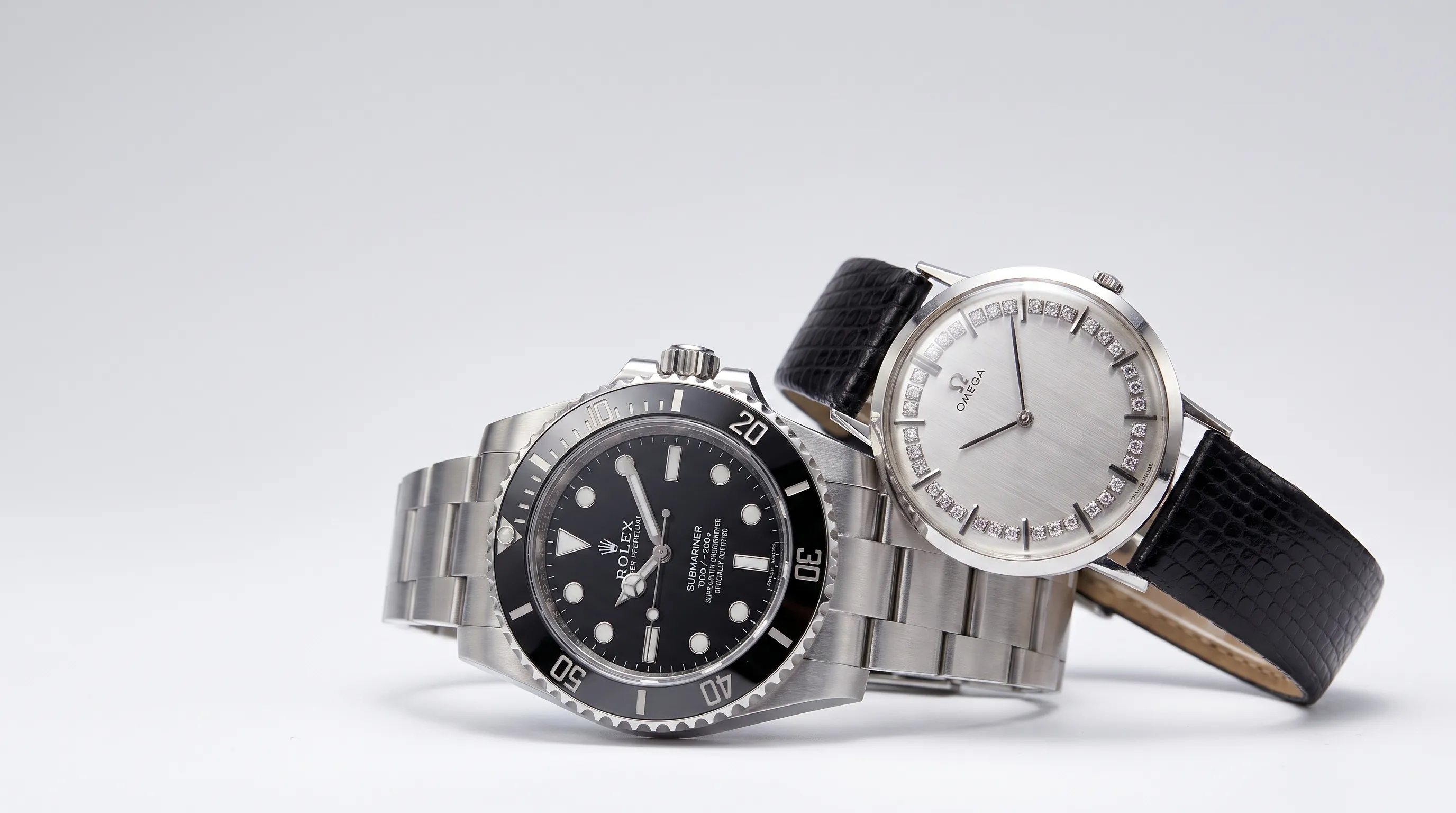 Two different luxury watches arranged side by side on a clean gradient studio backdrop, one stainless steel dive watch and one dress watch, both angled toward the camera as if being compared before purchase. The frame is medium-wide and balanced, with one watch slightly forward in the foreground and the other set back to create depth, leaving clear negative space in the top-left.