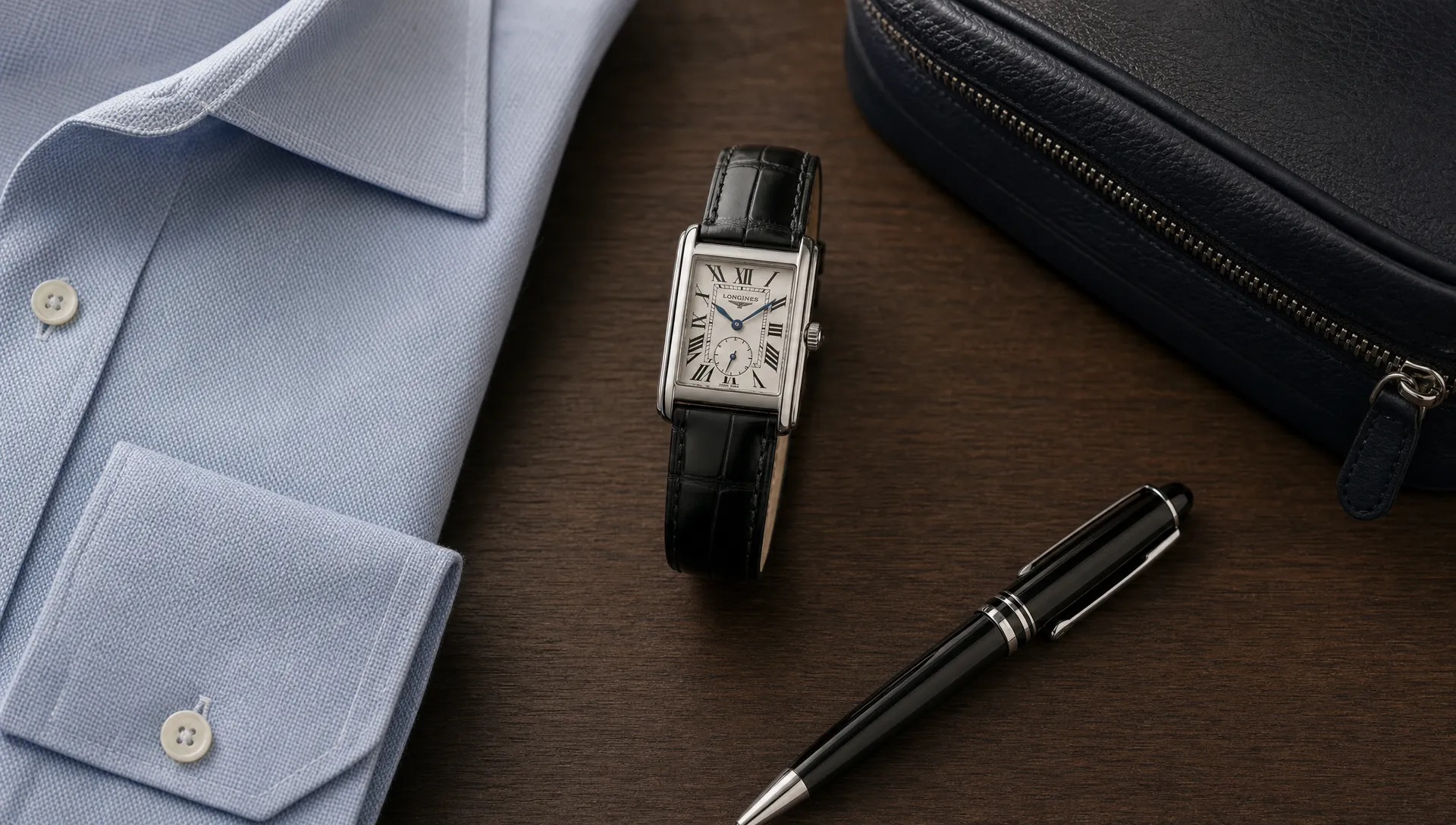 A rectangular dress watch on a leather strap sits beside a folded shirt, a pen and a travel pouch, emphasizing its office-ready proportions.