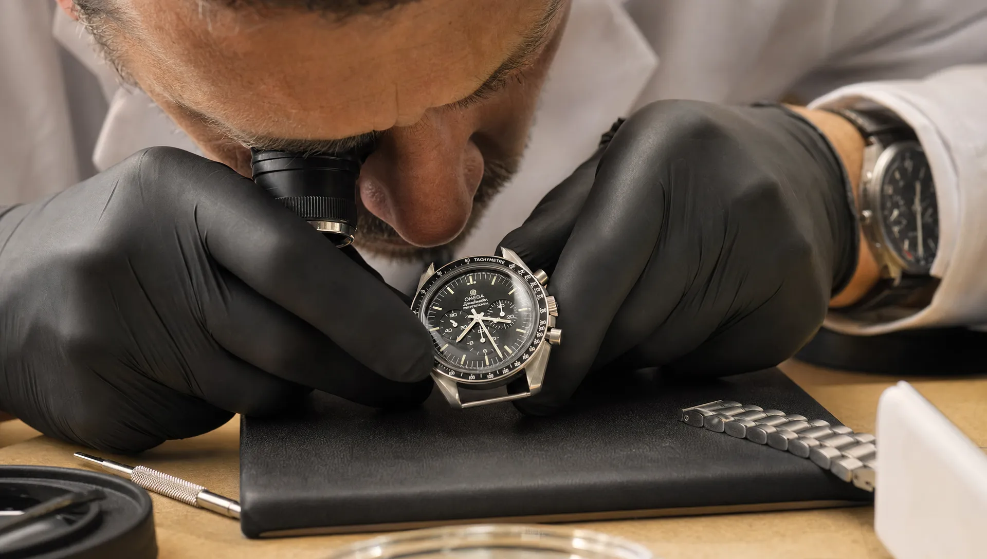 A watchmaker examines the lug edges and crystal of a pre-owned Omega Speedmaster on a padded work mat, with bracelet links nearby.