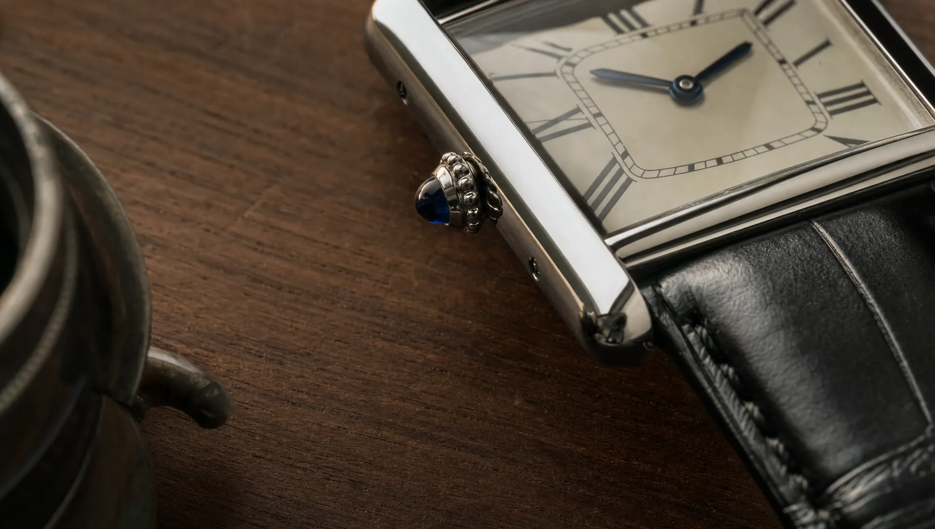 A vintage Cartier Tank case corner and cabochon crown sit beside a loupe on a watchmaker’s bench.