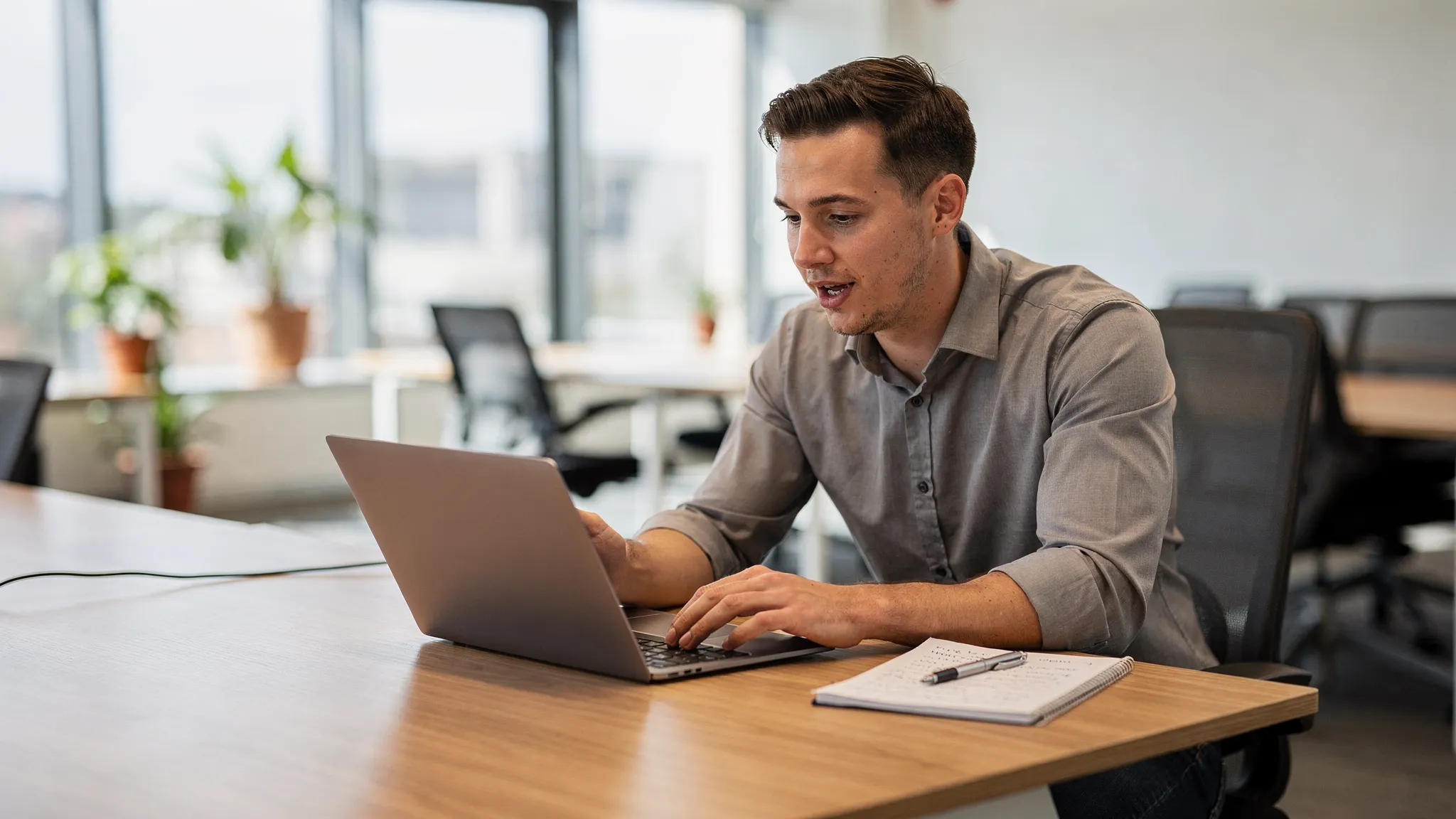 A sales development rep practicing an AI roleplay simulation on a laptop in a modern startup office, seated at a desk with natural light. The laptop screen faces the person, nothing visible behind it. The rep speaks while reading feedback prompts and notes beside the screen.