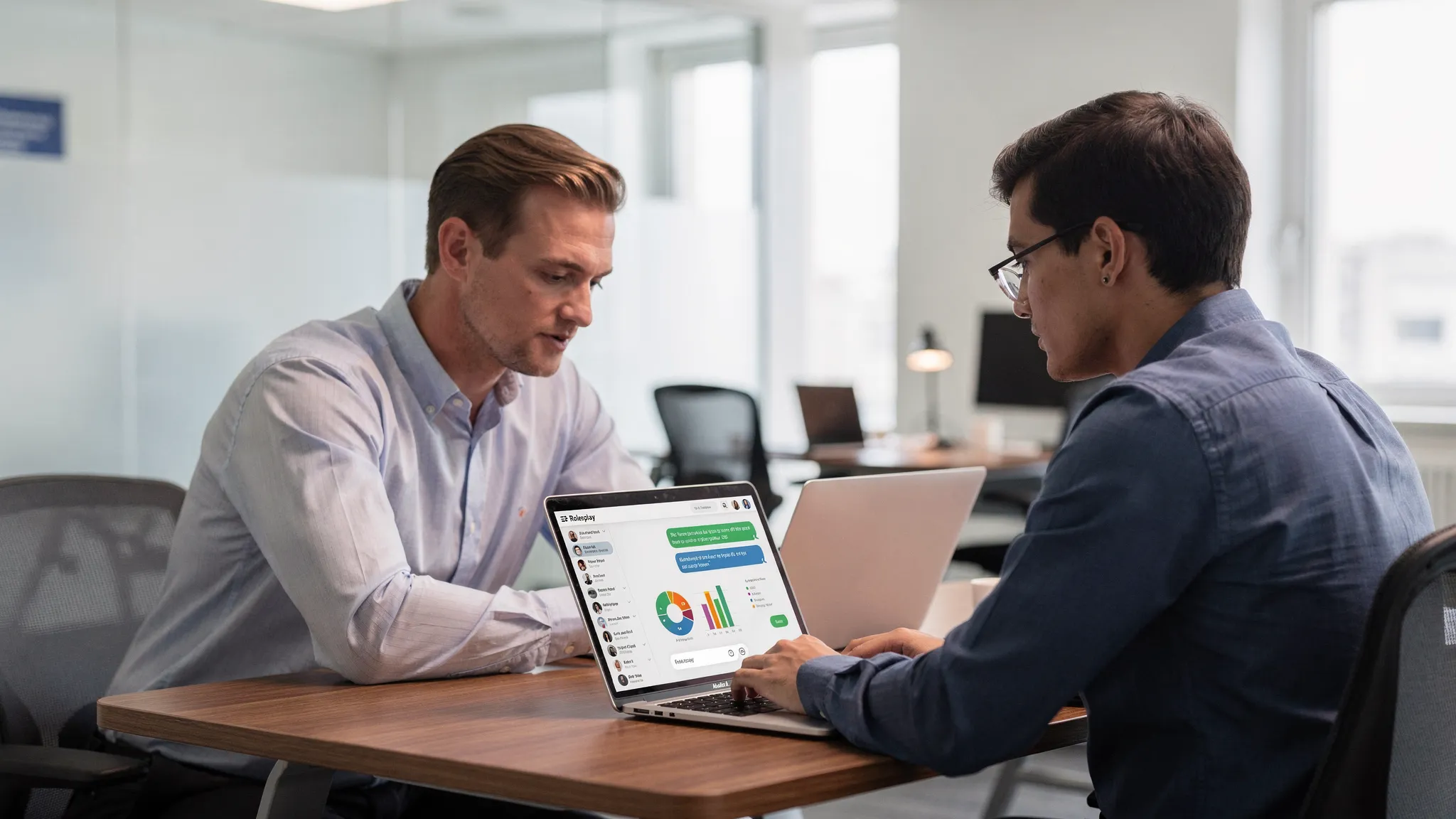A sales manager reviewing a dashboard while a new rep practices an AI roleplay on a laptop, both seated at a table in a professional office setting. The laptop screen faces the rep and shows a roleplay chat interface.