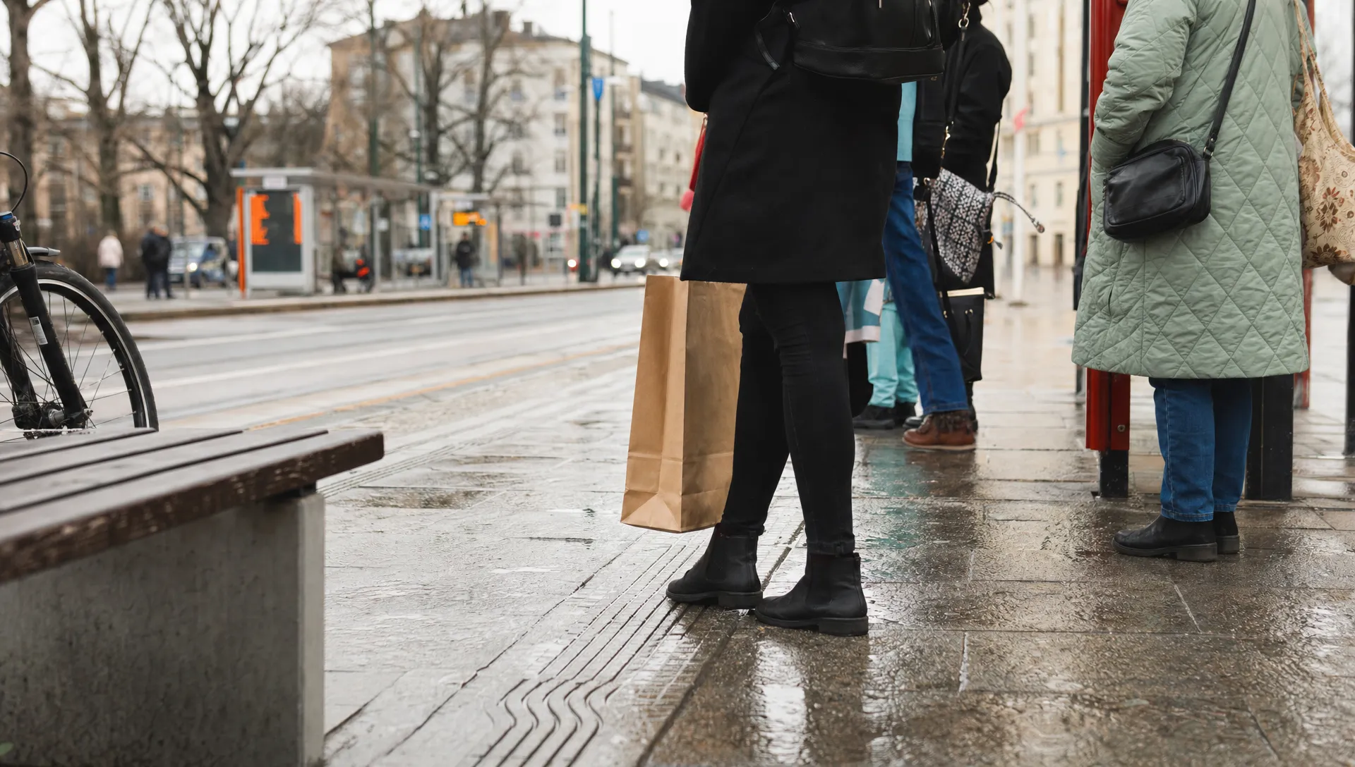 Commuters wait at a Warsaw tram stop beside wet paving and a bicycle, part of daily life in the city.