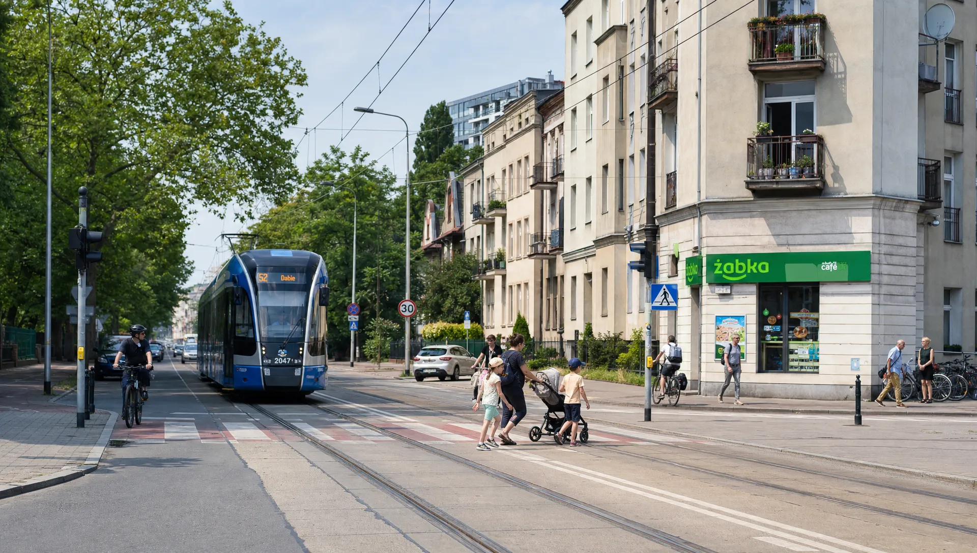 A walkable Polish neighborhood with tram tracks, apartment buildings, trees, cyclists and families crossing near a local shop.