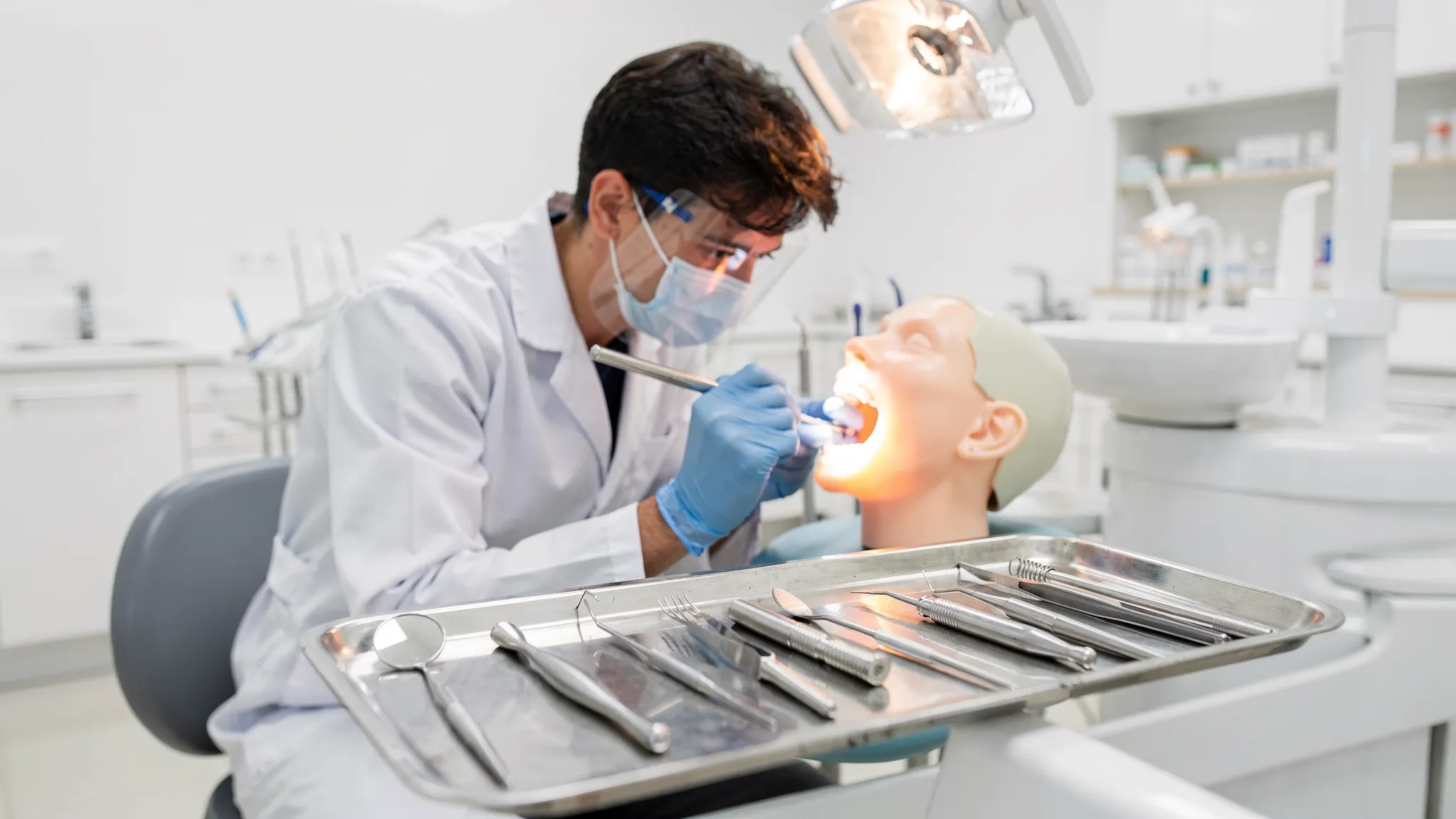 A student in a dental college simulation lab wearing gloves and a lab coat, practicing on a dental mannequin with basic dental instruments arranged on a tray.