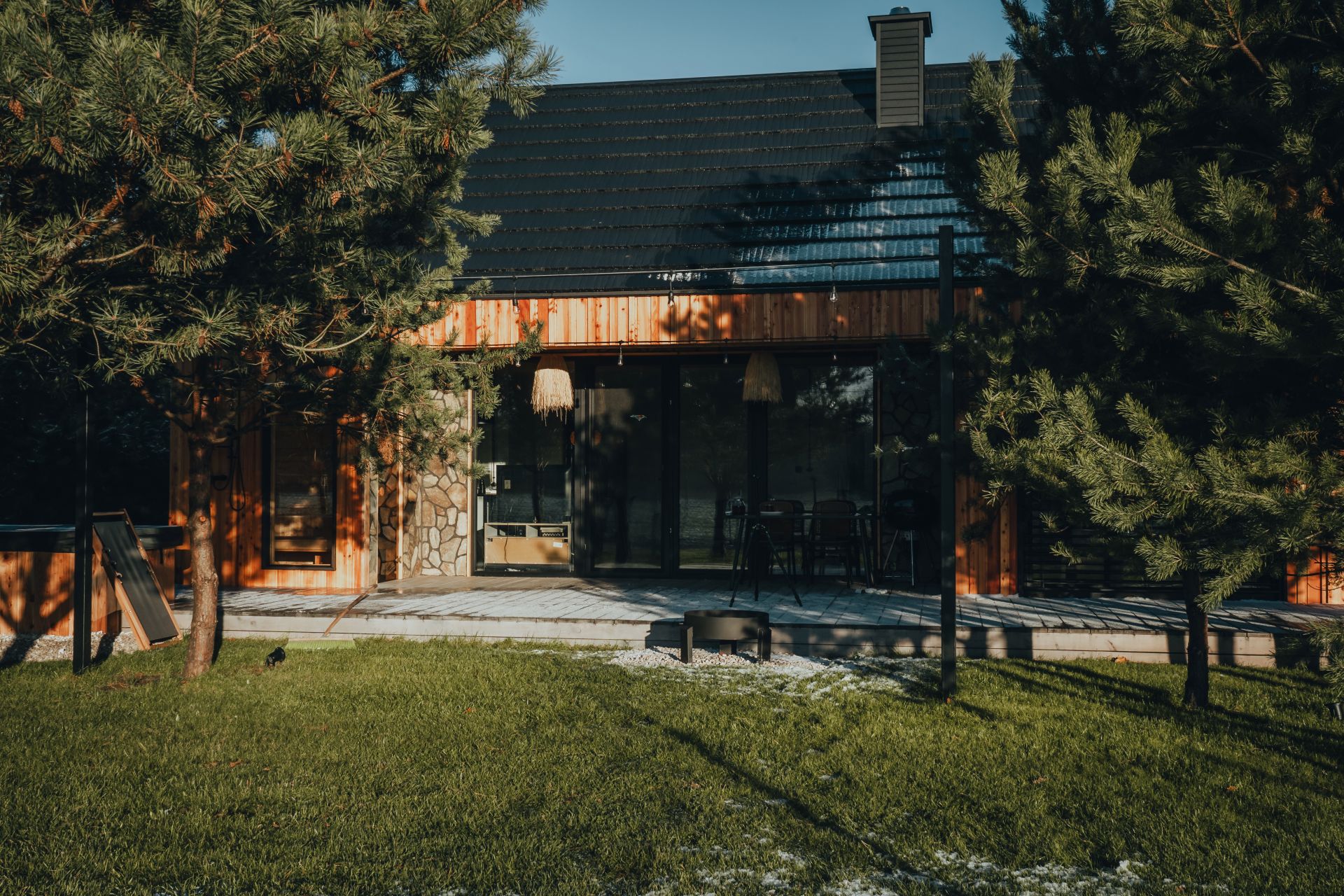 Landscape cover showing a relaxed weekend morning outside a cottage: a pair of bicycles leaned near the terrace, a coffee cup on the table, and a dog resting on the fenced lawn while the wooden house and tree line fill the background. Use a wide eye-level view that feels like ordinary use of the place rather than an arrival scene.
