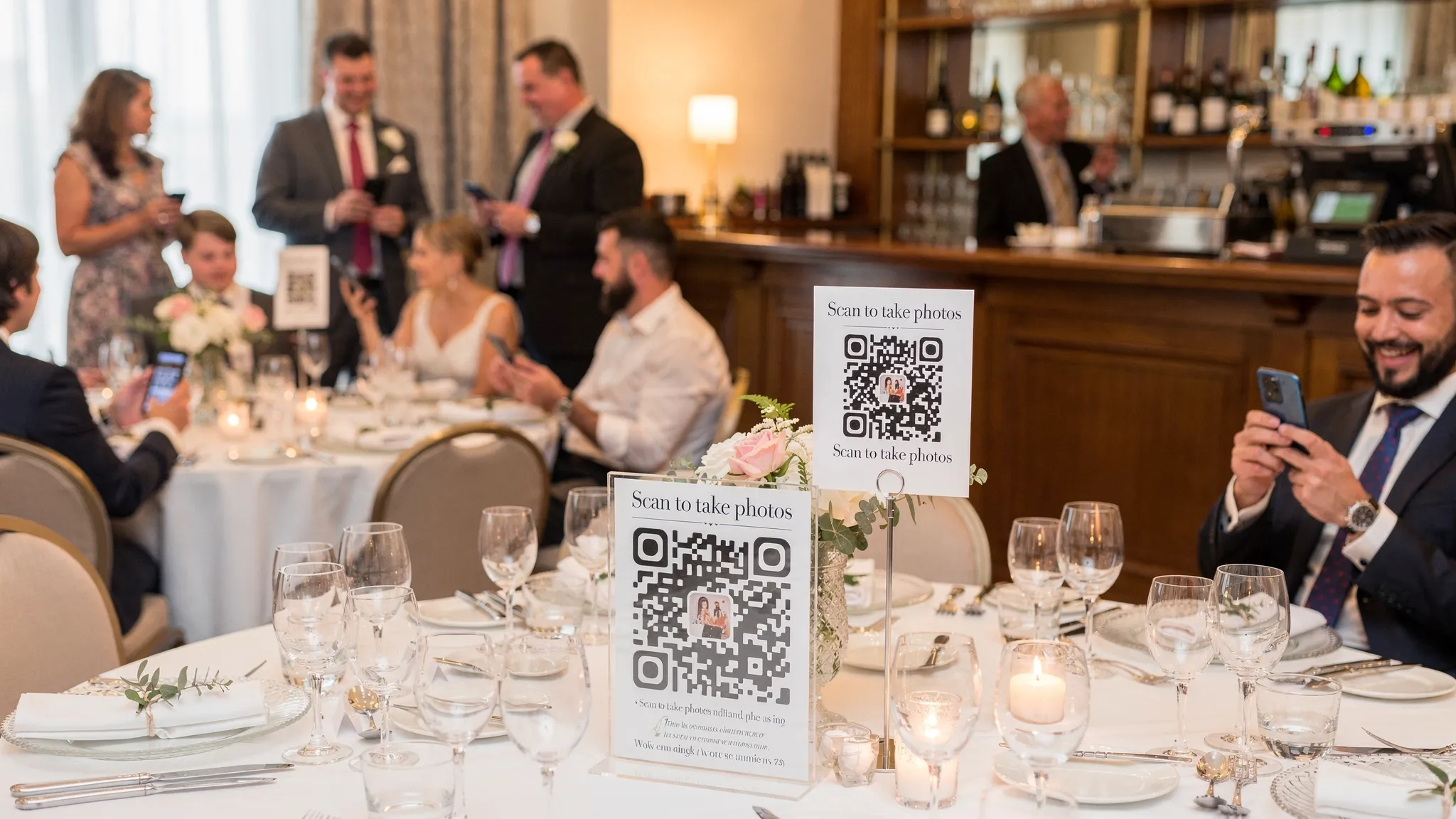 Event signage and table tent cards displaying a QR code labeled “Scan to take photos,” placed near a bar and on dinner tables at a wedding reception, with guests casually scanning using their phones.