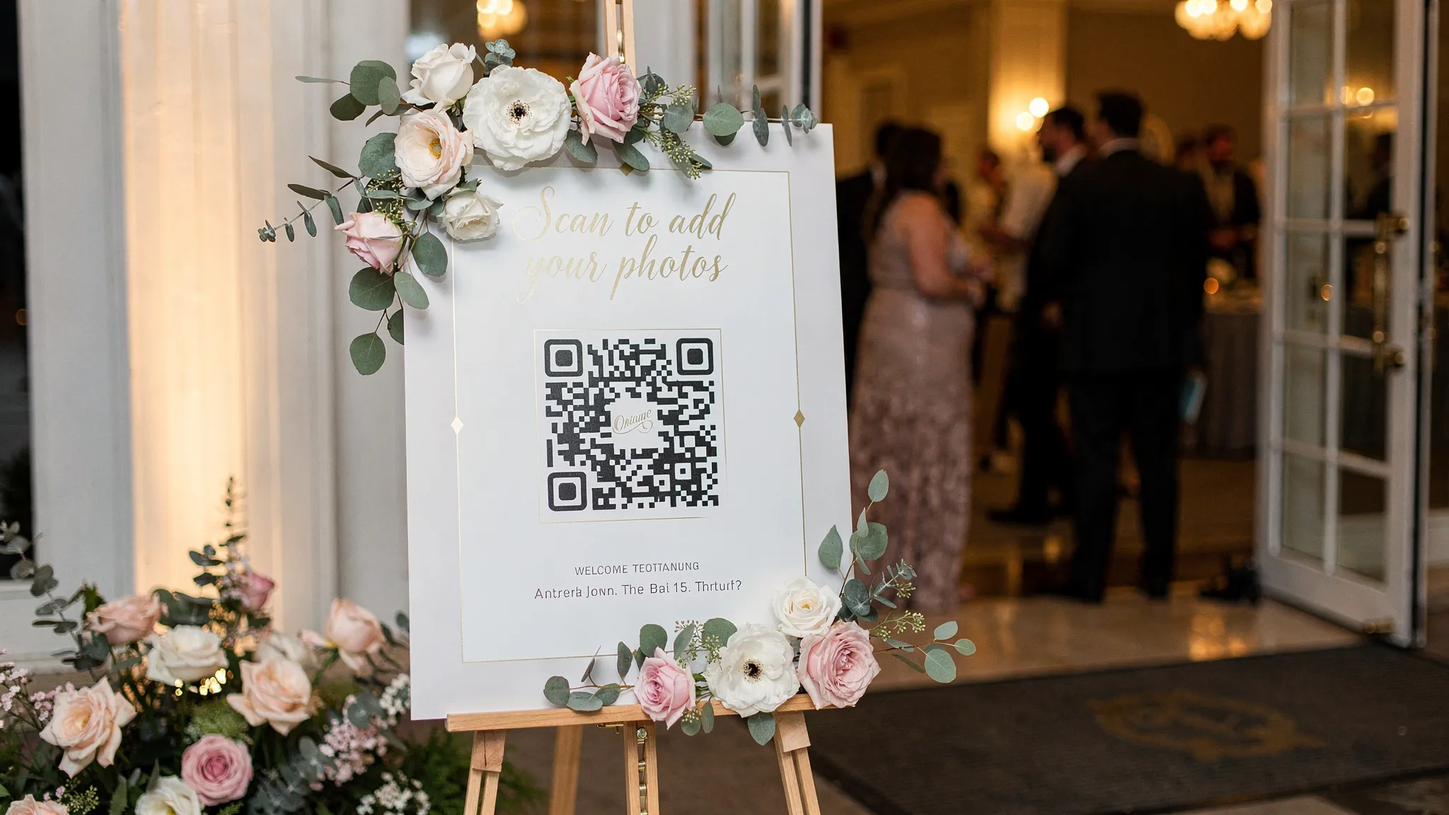A wedding reception welcome sign on an easel featuring a large QR code and a short call to action like “Scan to add your photos,” placed near the venue entrance with floral decor and guests arriving in the background.