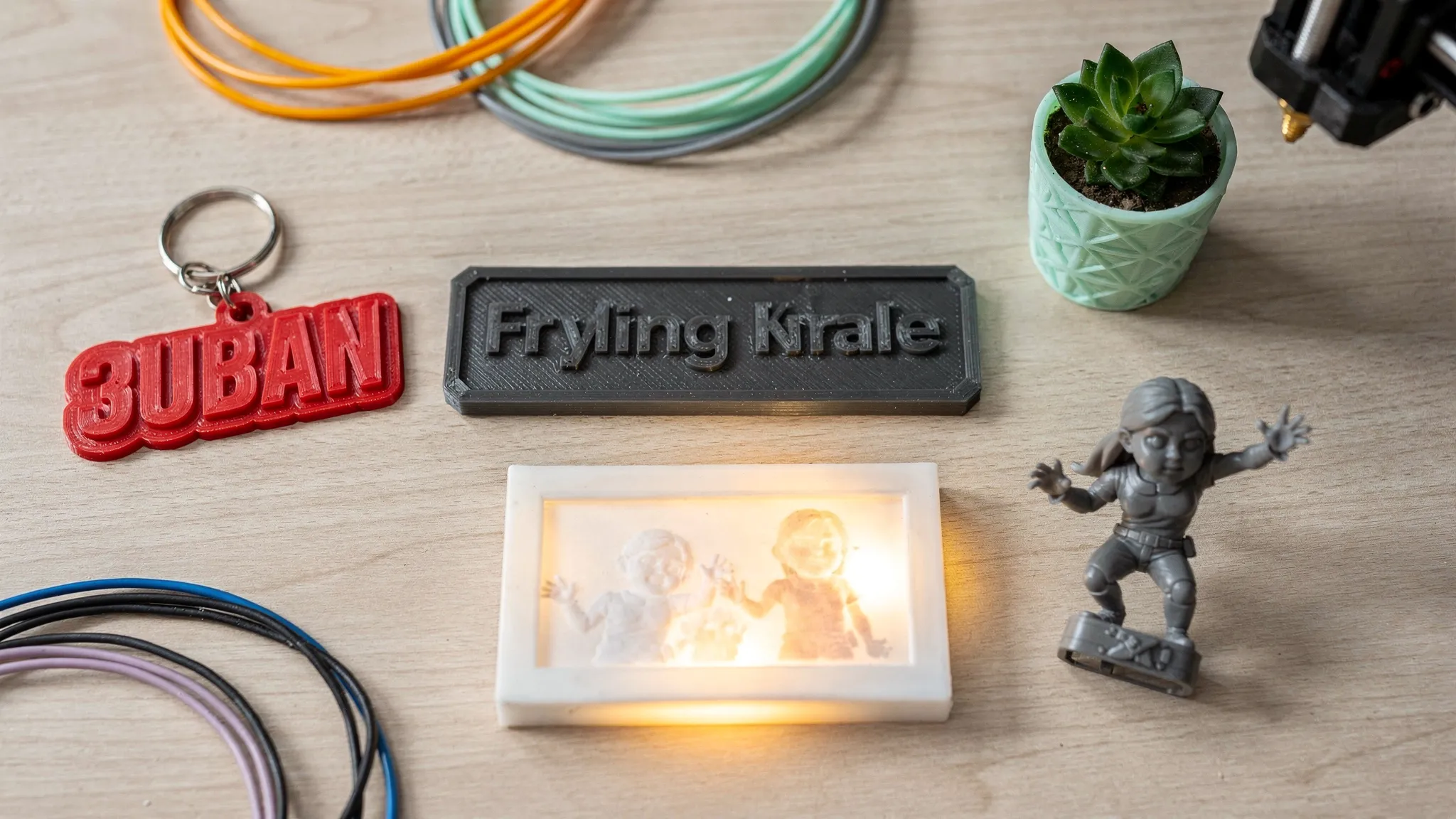 A flat lay collage of 3D printed gift ideas on a table: a personalized keychain, a custom name plate, a small plant pot, a lithophane night light, and a mini figurine, all in different colors and textures.