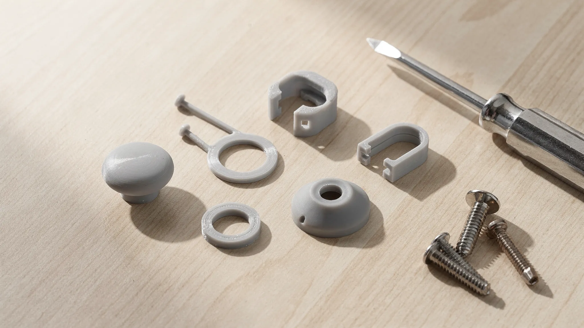 A neatly arranged set of small 3D printed household fix parts on a workbench, including a knob, a clip, a spacer ring, a cable strain relief, and a rubbery foot pad, shown next to a screwdriver and a few screws.