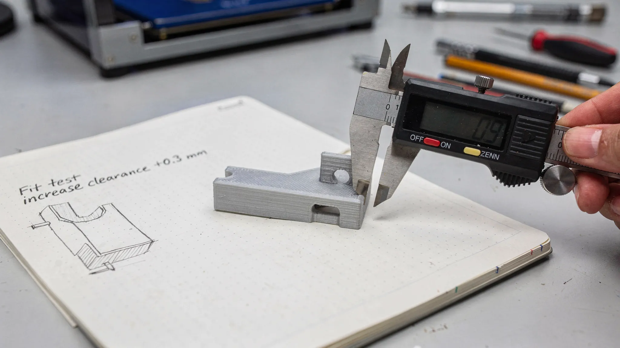 A photo-style scene of a workbench with a small 3D printed bracket, calipers measuring a feature, and a notebook showing a simple fit test note like “increase clearance +0.3 mm.” The focus is on iteration and measurement.