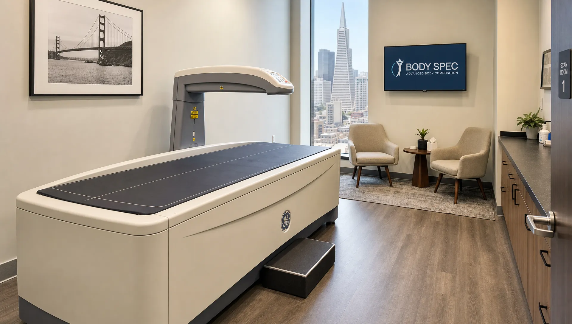 Landscape interior of a professional body composition scan room in San Francisco, with the DEXA scanner table leading toward the camera, the scanner arm beside it, and a small consultation corner with chairs and a wall monitor in the background; the room is empty and the scan setup is the focus. Wide eye-level composition, calm clinical setting.
