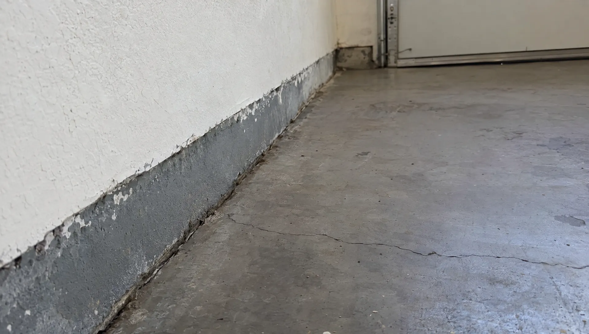 A close view of an empty garage slab meeting the wall, with a small crack and old paint edge visible for the installer to assess before epoxy floor coating.