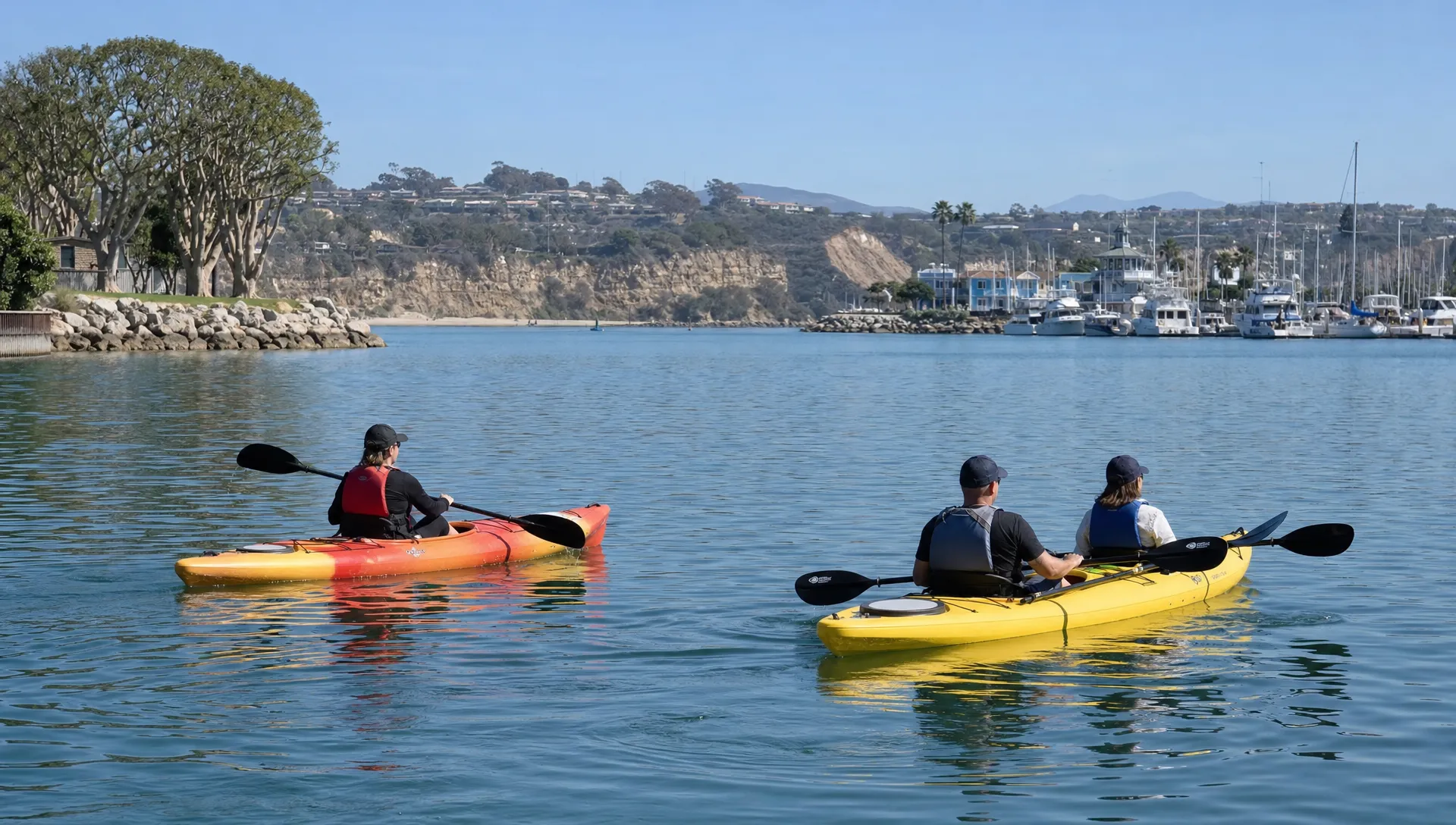 Two kayaks move across calm Dana Point Harbor water, one with a solo paddler and one with two paddlers sharing the boat.