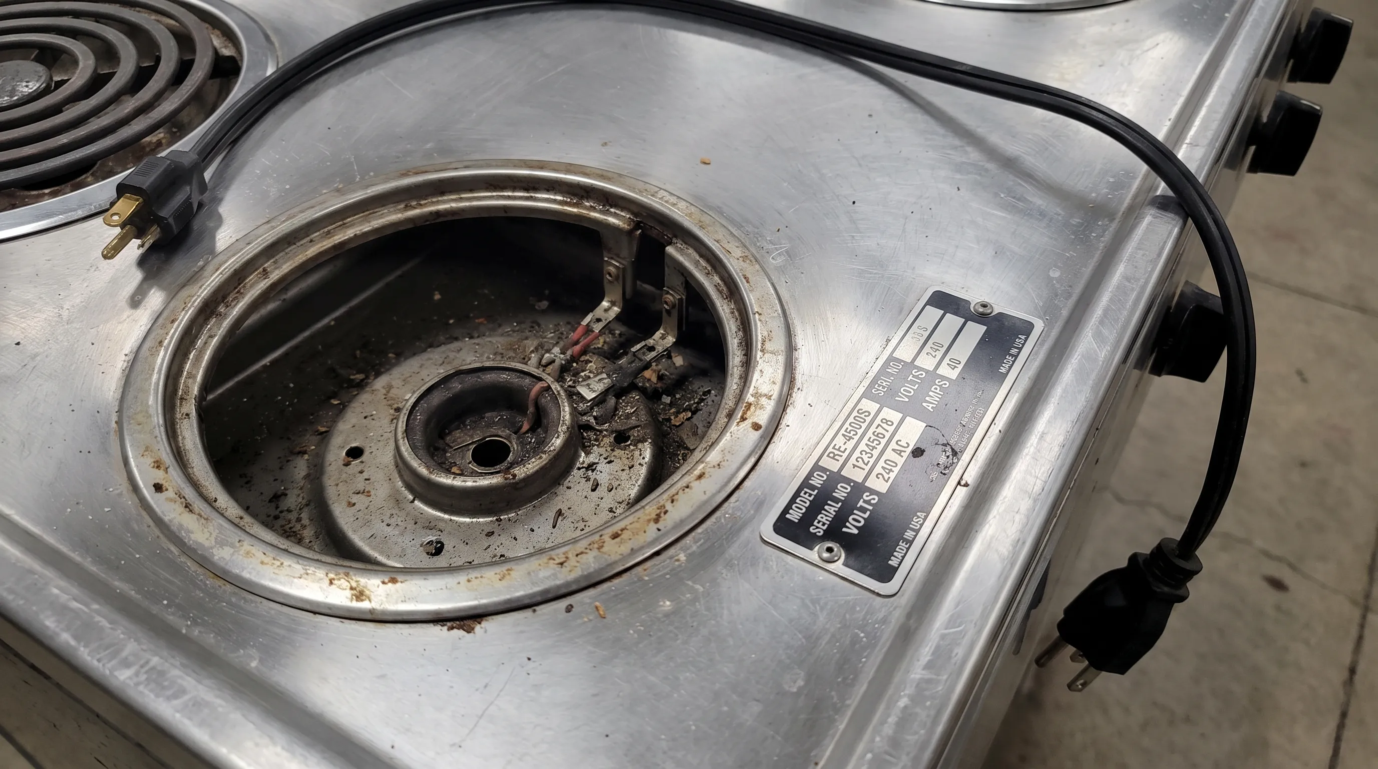 A close view of a disconnected electric range with its coil element removed, showing the burner receptacle and nearby model label during inspection.