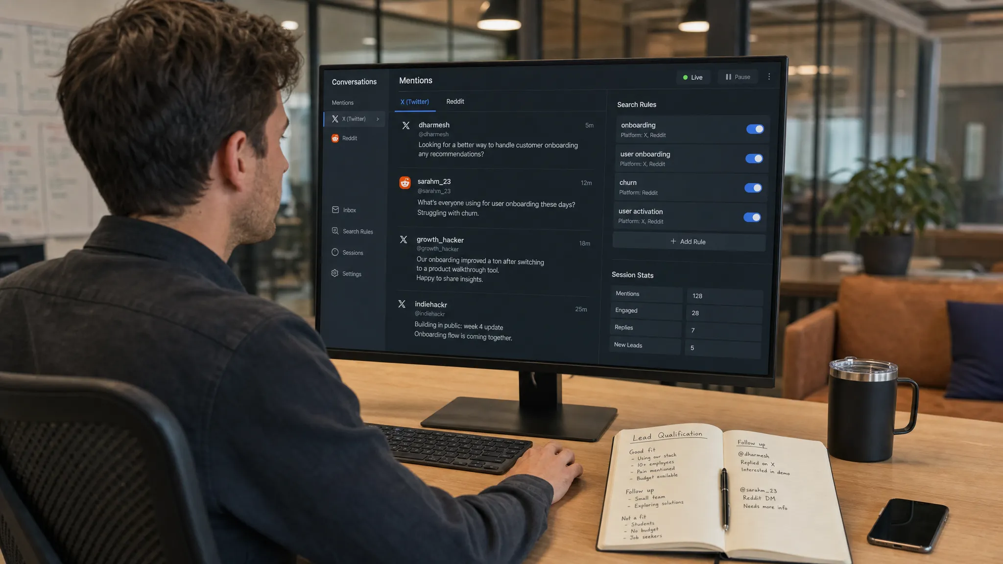 A founder at a desk reviewing a real-time conversation dashboard split between X and Reddit mentions, with an inbox of relevant posts, search rules, and session stats on a monitor facing the camera, plus a notebook with lead qualification notes and a phone beside it in a wide office scene.