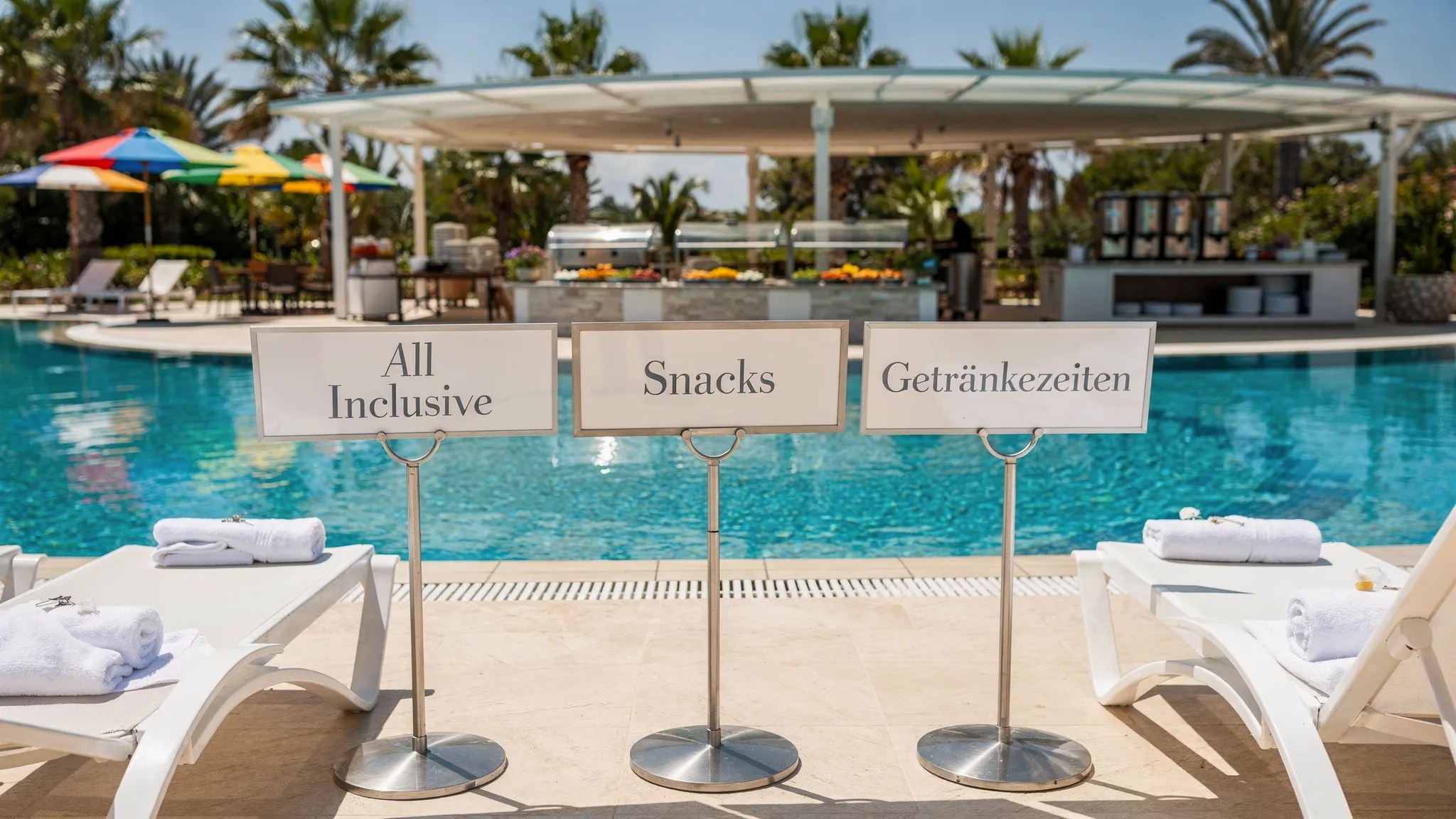 Eine sonnige Resort-Szene mit Pool, Liegen und einem Buffetbereich im Hintergrund. Im Vordergrund stehen Schilder mit &bdquo;All Inclusive&ldquo;, &bdquo;Snacks&ldquo; und &bdquo;Getr&auml;nkezeiten&ldquo;, um die typischen Inklusivleistungen und Zeitfenster zu visualisieren.
