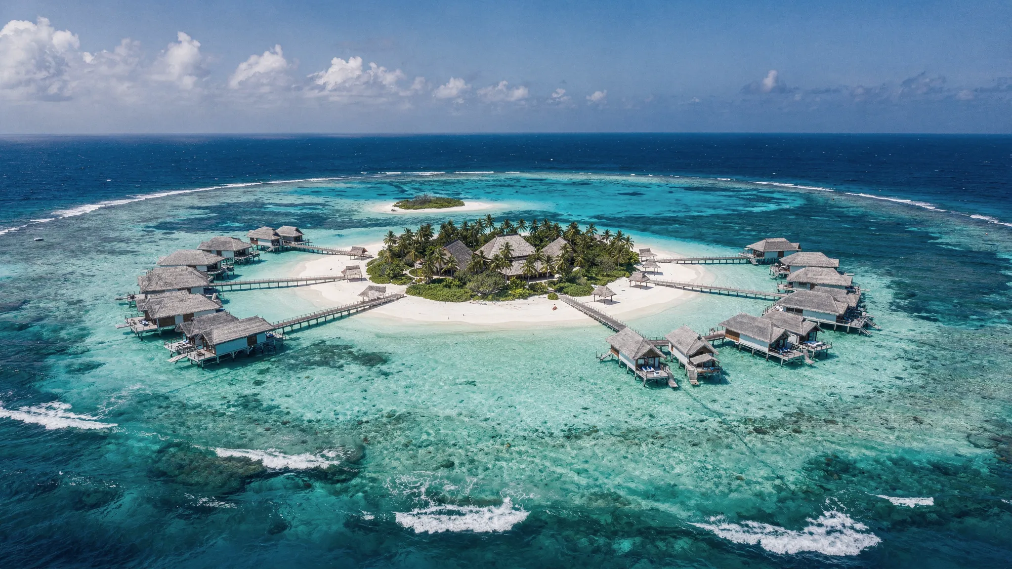 Luftaufnahme einer kleinen Malediveninsel mit Stegen zu Overwater-Villen, hellem Sandstrand, türkisfarbenem Lagunenwasser und tieferem Blau am Außenriff.
