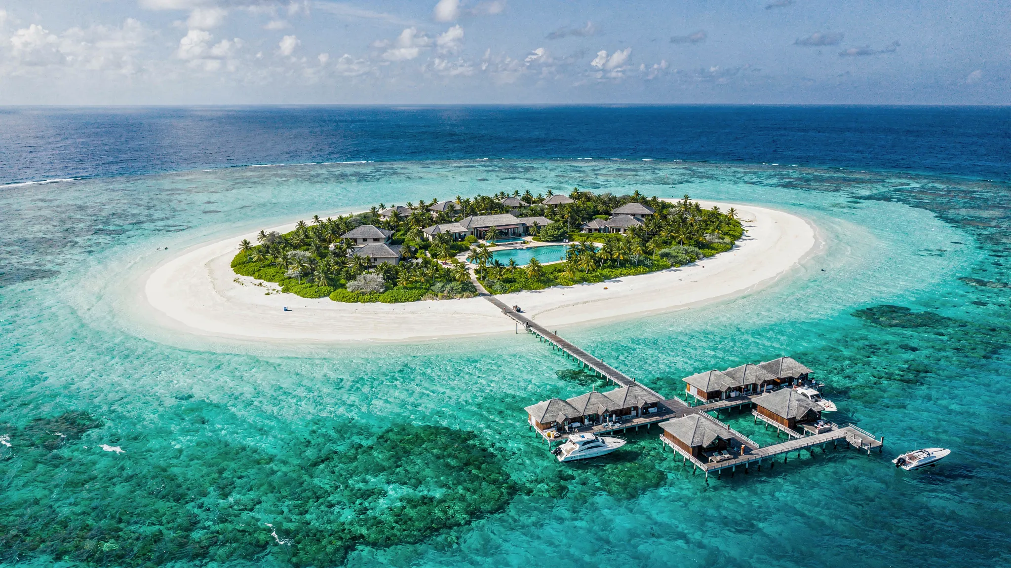 Luftaufnahme einer grünen Malediven-Insel mit umlaufendem weißen Sandstrand, türkisfarbener Lagune und einem Resort mit Stegen und Bootsanleger.