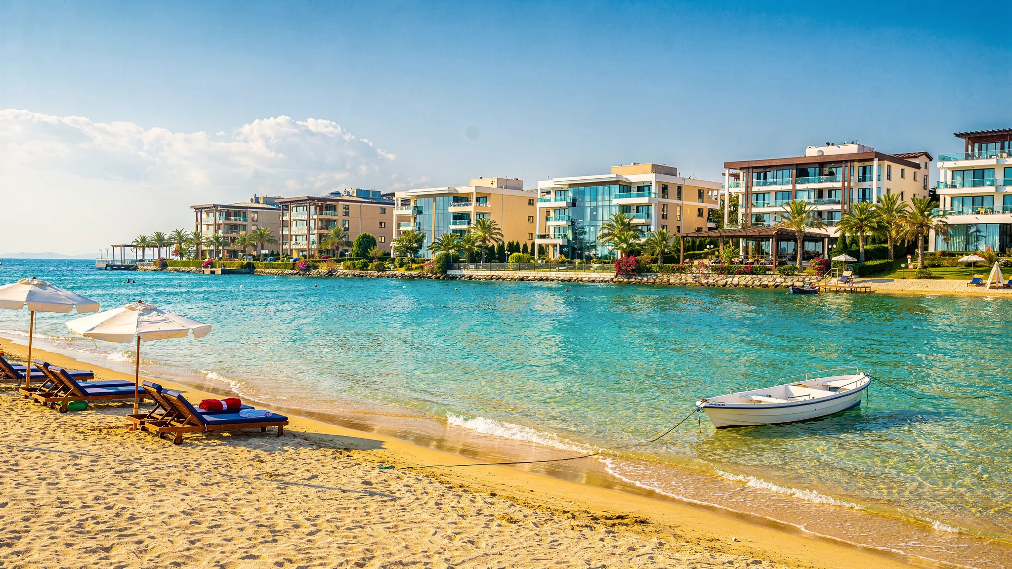 Ein sonniger Sandstrand an der Türkischen Riviera mit türkisfarbenem Wasser, im Hintergrund moderne Hotelresorts und Palmen, dazu eine kleine Szene mit Liegen und einem Boot in Küstennähe.