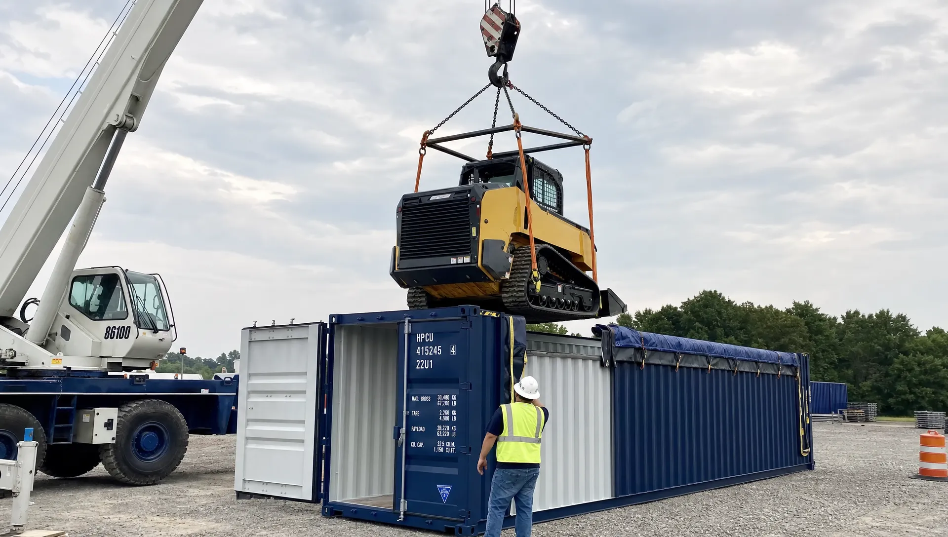 A 20ft open top container with its tarp rolled back, roof bows removed, and a crane lowering a large piece of construction equipment into the cargo container from above on a gravel job site.
