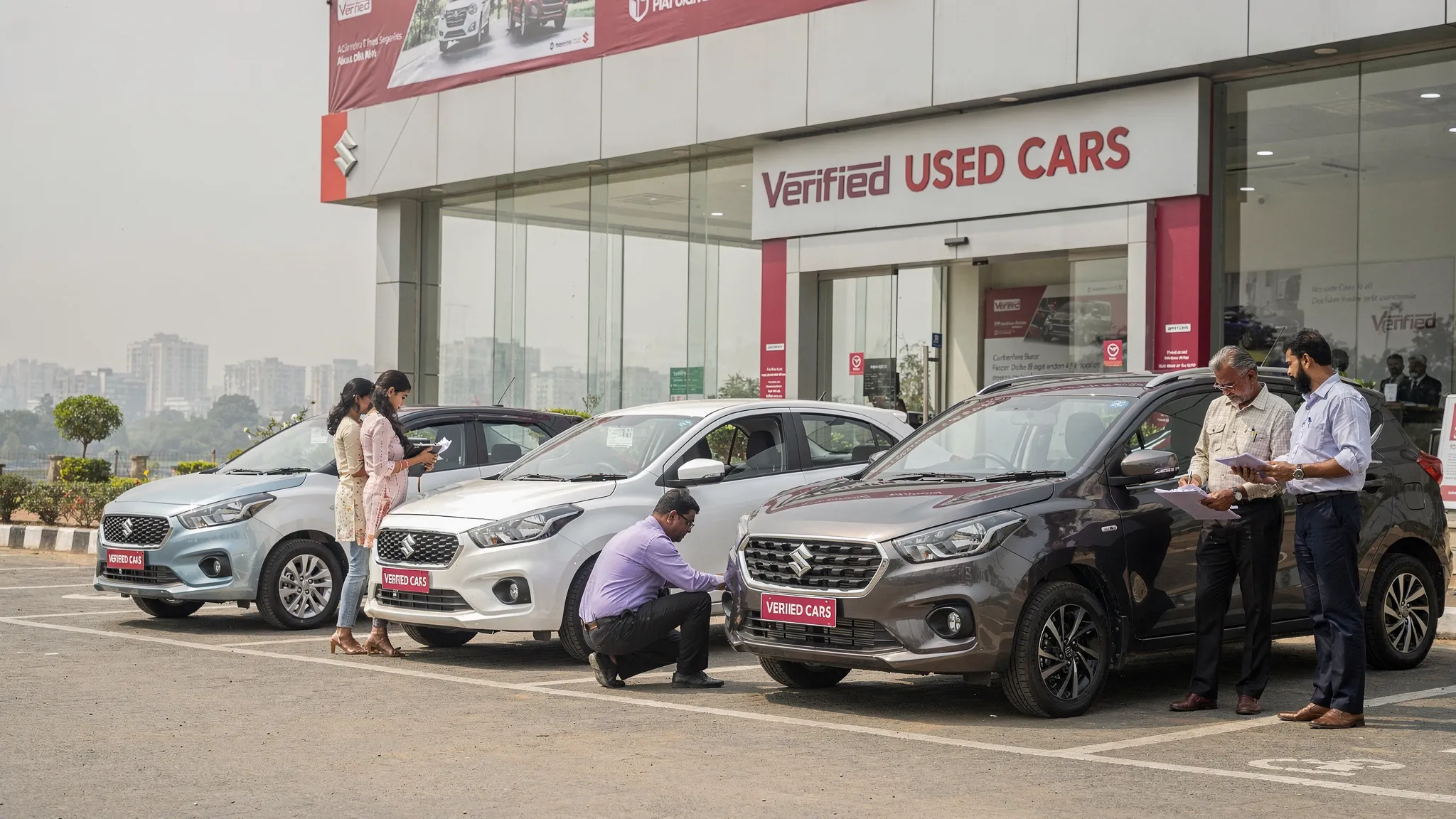 A row of clean used Maruti cars parked at a verified dealership in Delhi NCR, including a hatchback, sedan, compact SUV, and MPV, with buyers inspecting documents and vehicle condition.