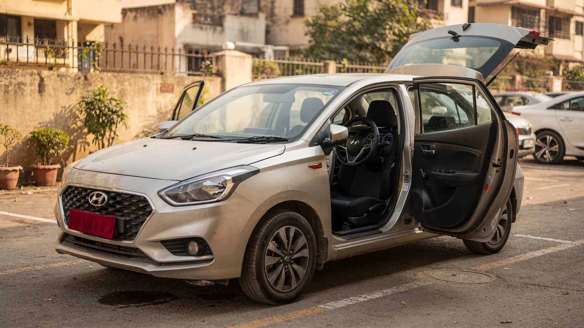 A clean used car parked in a shaded Delhi NCR residential parking area, photographed from the front three-quarter angle with visible tyres, clear body panels, open interior door, dashboard, odometer area, and boot space ready for a buyer inspection.