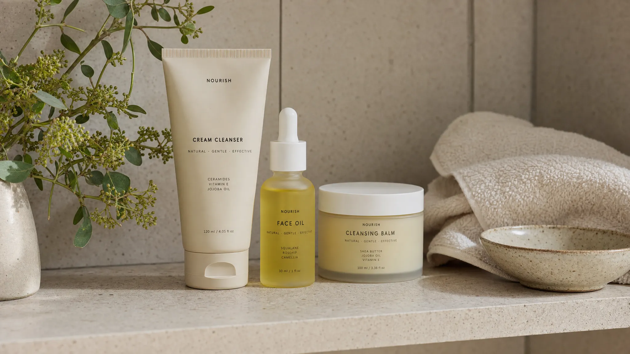 A close-up view of a bathroom shelf with a cleanser, face oil, and balm arranged beside a soft cotton towel, fresh botanical sprigs, and a ceramic bowl.