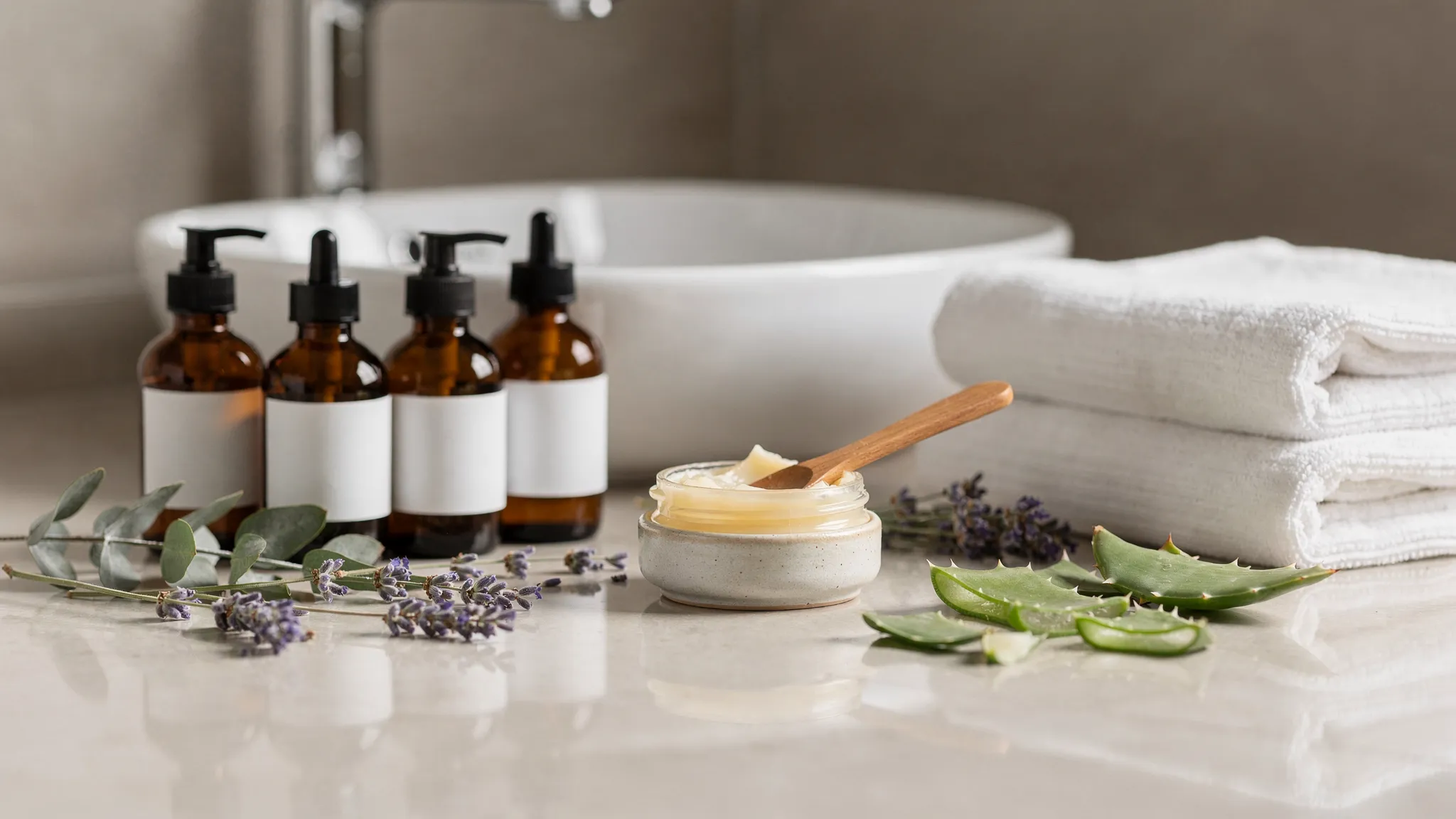 A calm apothecary-style bathroom counter with amber glass bottles, a small ceramic jar of balm, folded cotton towels, and fresh botanical ingredients arranged beside a sink.