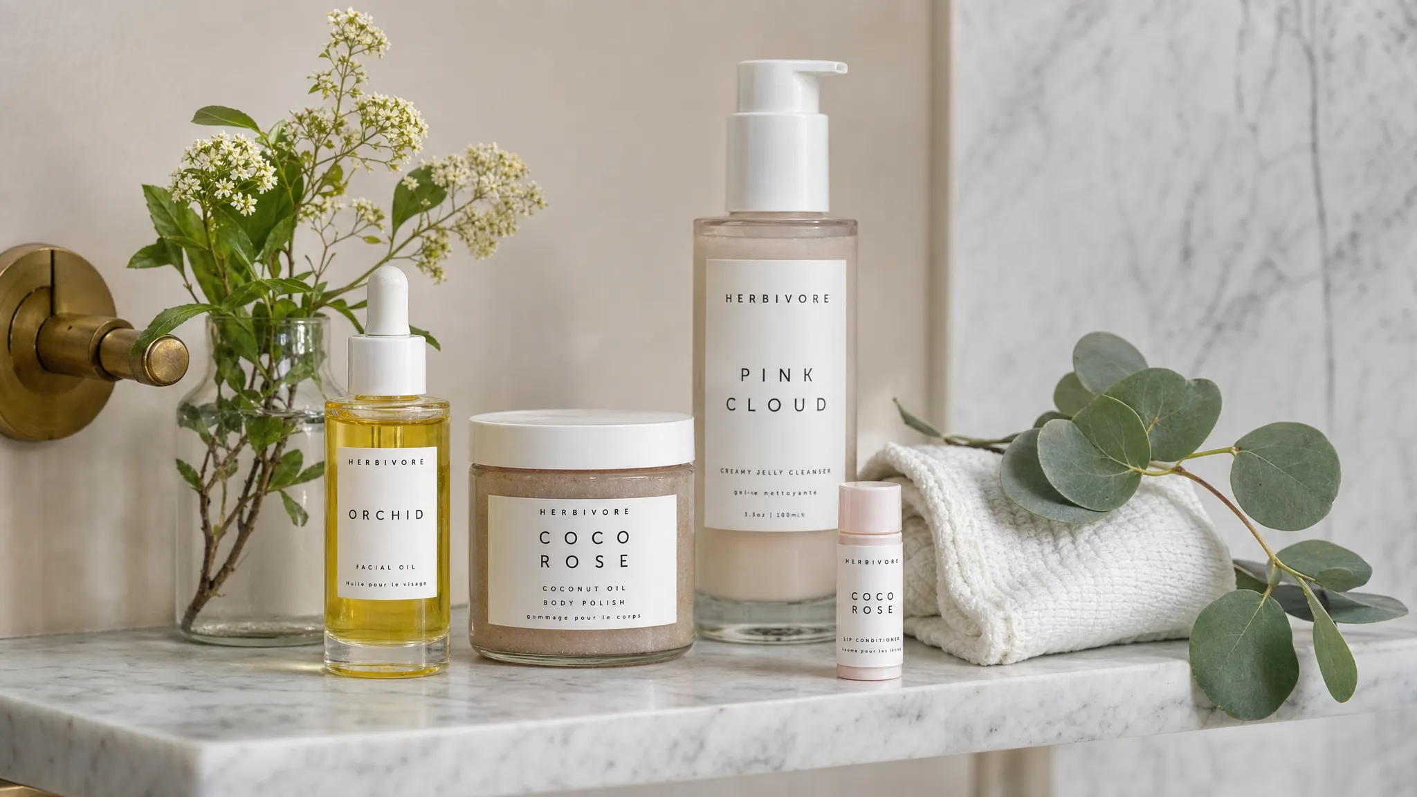 A calm bathroom shelf with a small group of natural skincare products, including a glass oil bottle, a balm jar, a cleanser, a lip balm, and a folded cotton towel beside fresh botanicals.