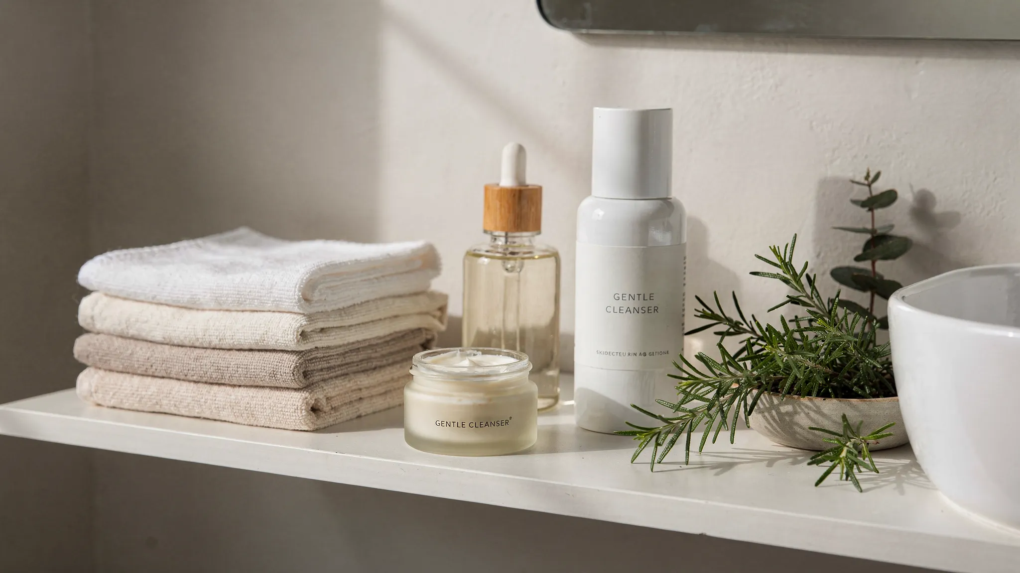 A calm bathroom shelf with botanical skincare essentials including a glass oil bottle, a balm jar, a gentle cleanser, folded cotton towels, and fresh herbs arranged beside a ceramic sink.