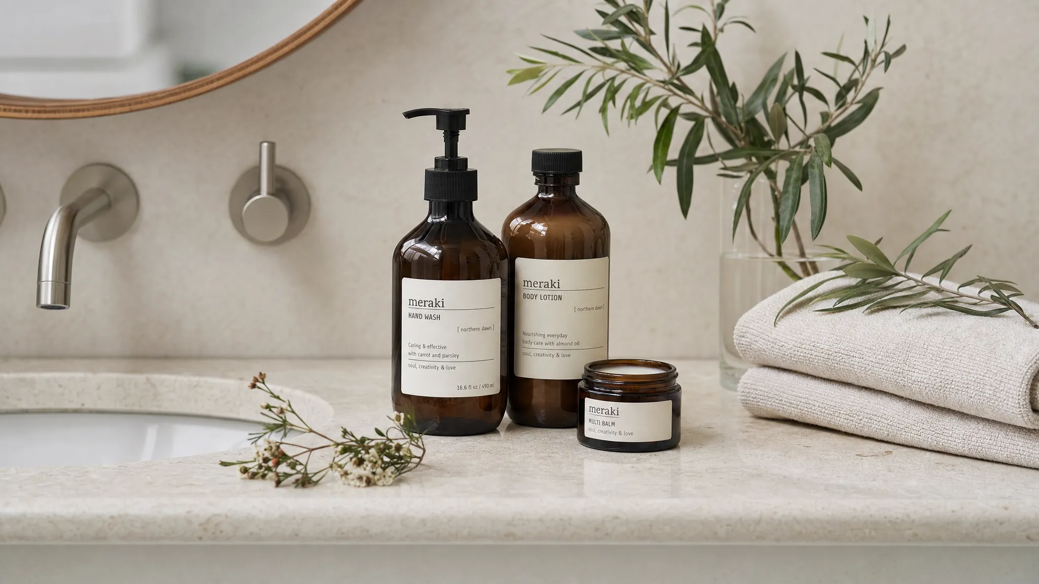 A simple bathroom sink scene with natural body care products, including amber glass bottles, a small balm jar, folded cotton towels, and fresh botanical sprigs on a stone counter.