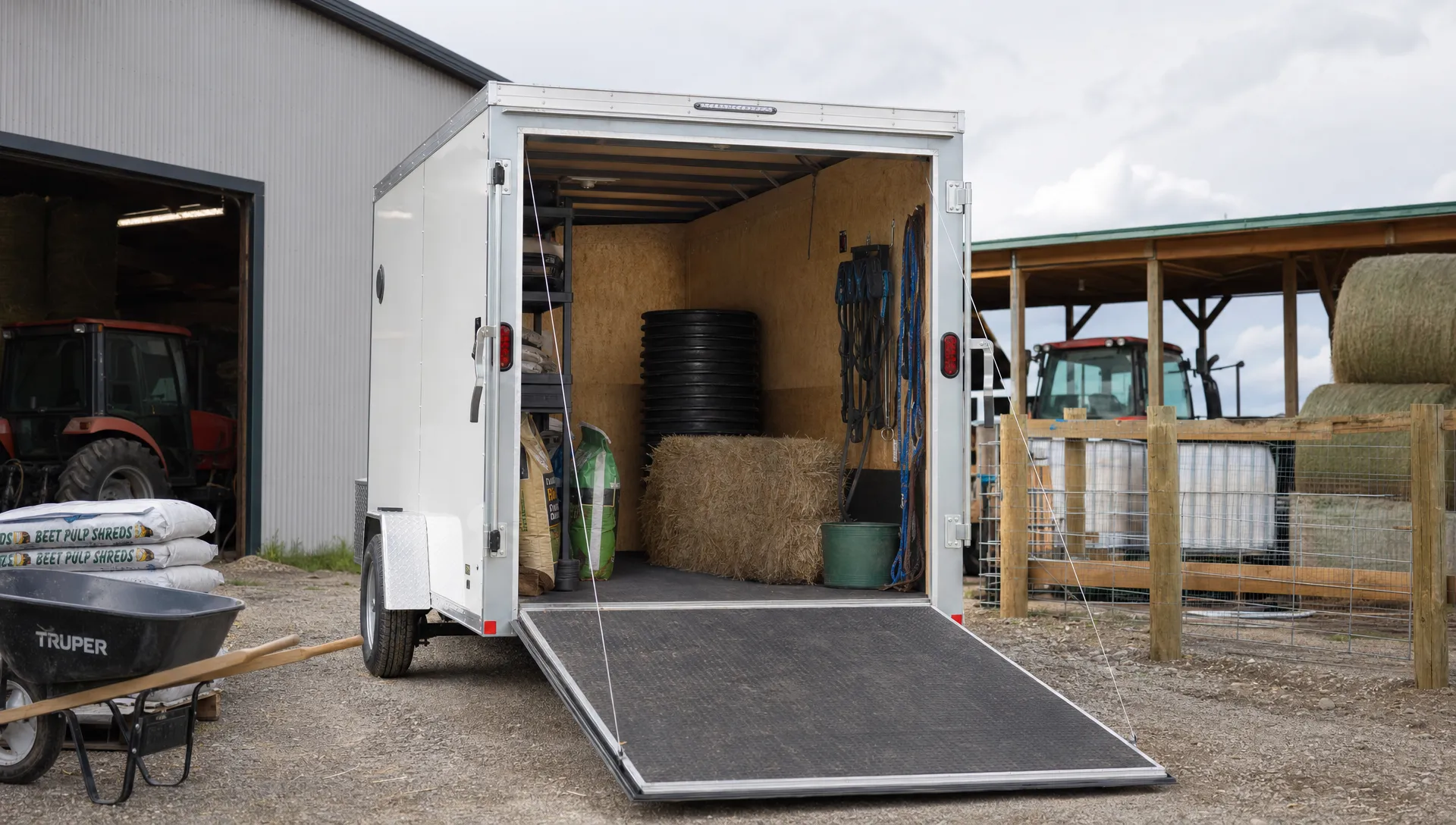 A box trailer sits open in a farmyard beside feed bags and fencing supplies.