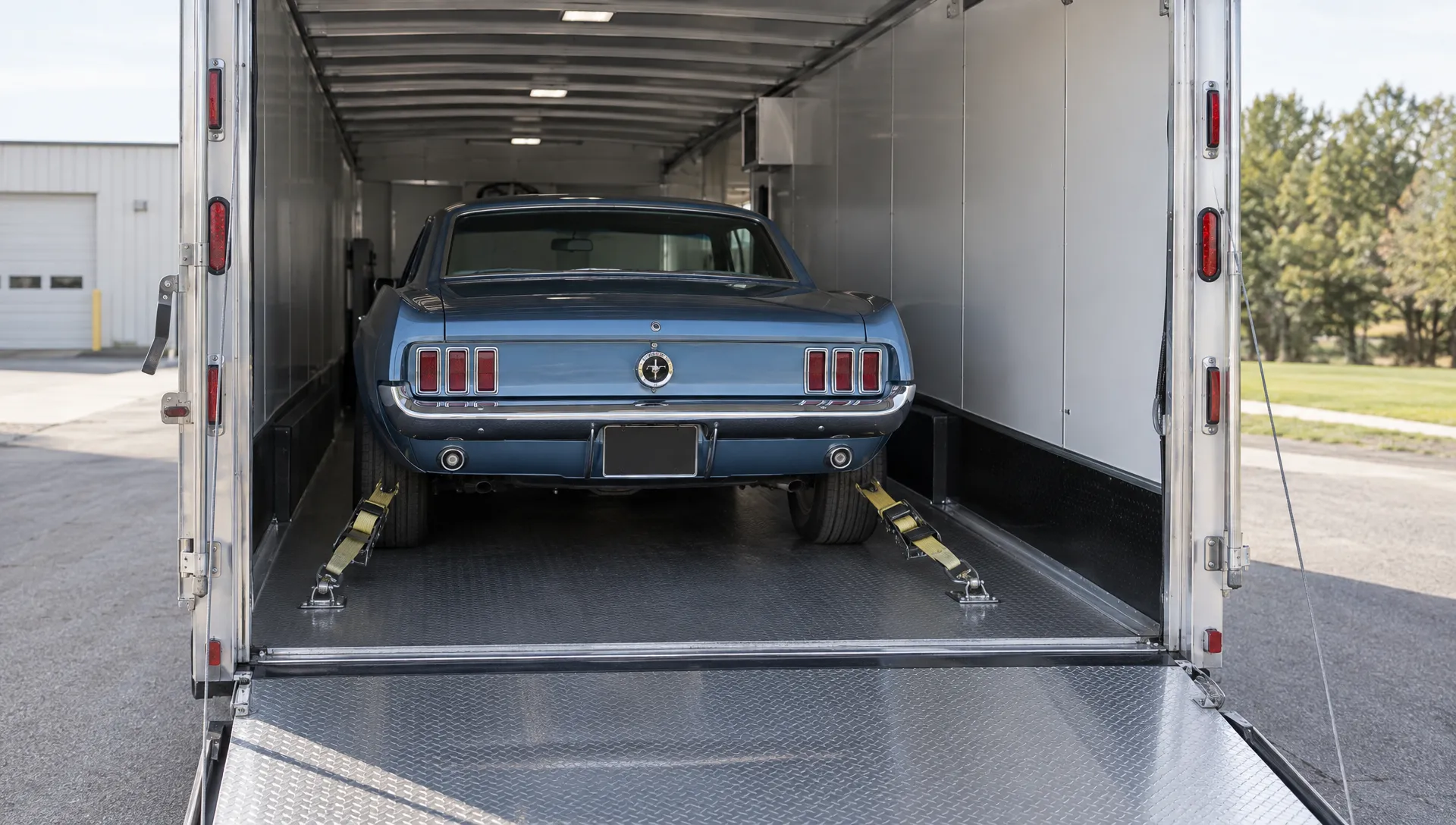 A classic coupe secured with wheel straps inside an enclosed car trailer, with clearance beside the vehicle and the loading ramp lowered onto level pavement.