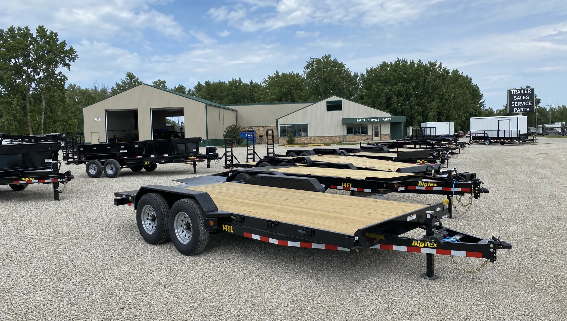 Tilt deck and equipment trailers sit in rows on a dealership lot beside the office and service bay. Tilt deck and equipment trailers sit in rows on a dealership lot beside the office and service bay.